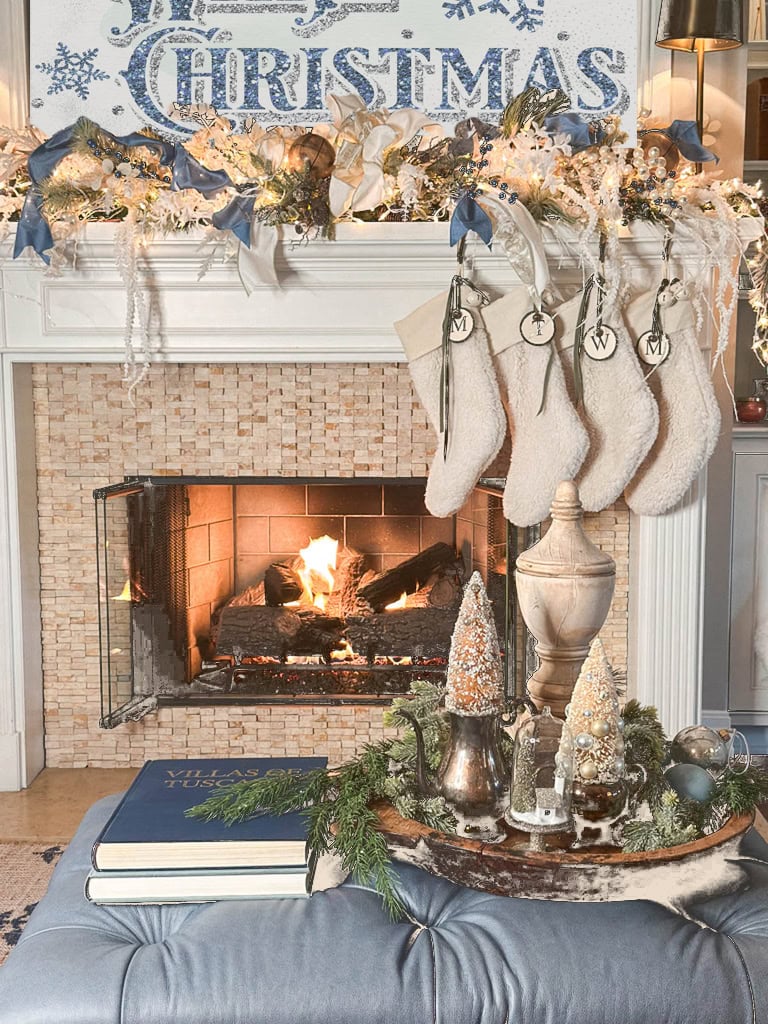 A cozy living room with a decorated Christmas mantel. Stockings hang above a lit fireplace, and a festive display with miniature trees and greenery adorns the coffee table. The word "Christmas" is displayed above the mantel.