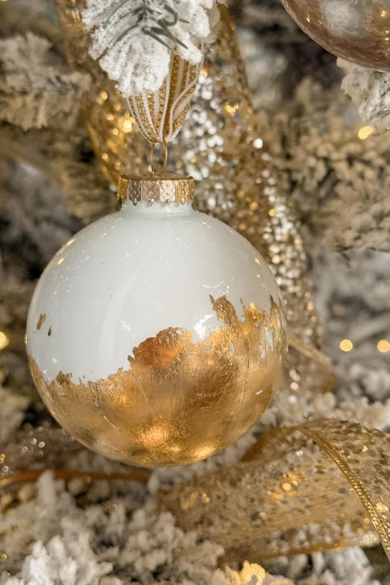 A white and gold ornament hangs on a Christmas tree adorned with snowy branches and shimmering gold ribbons. The ornament features a gold leaf pattern that adds a festive, elegant touch.