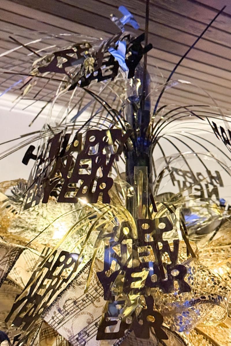 A festive New Year's centerpiece with shiny metallic "Happy New Year" decorations and streamers. The background is softly lit, highlighting the intricate details and celebratory theme.