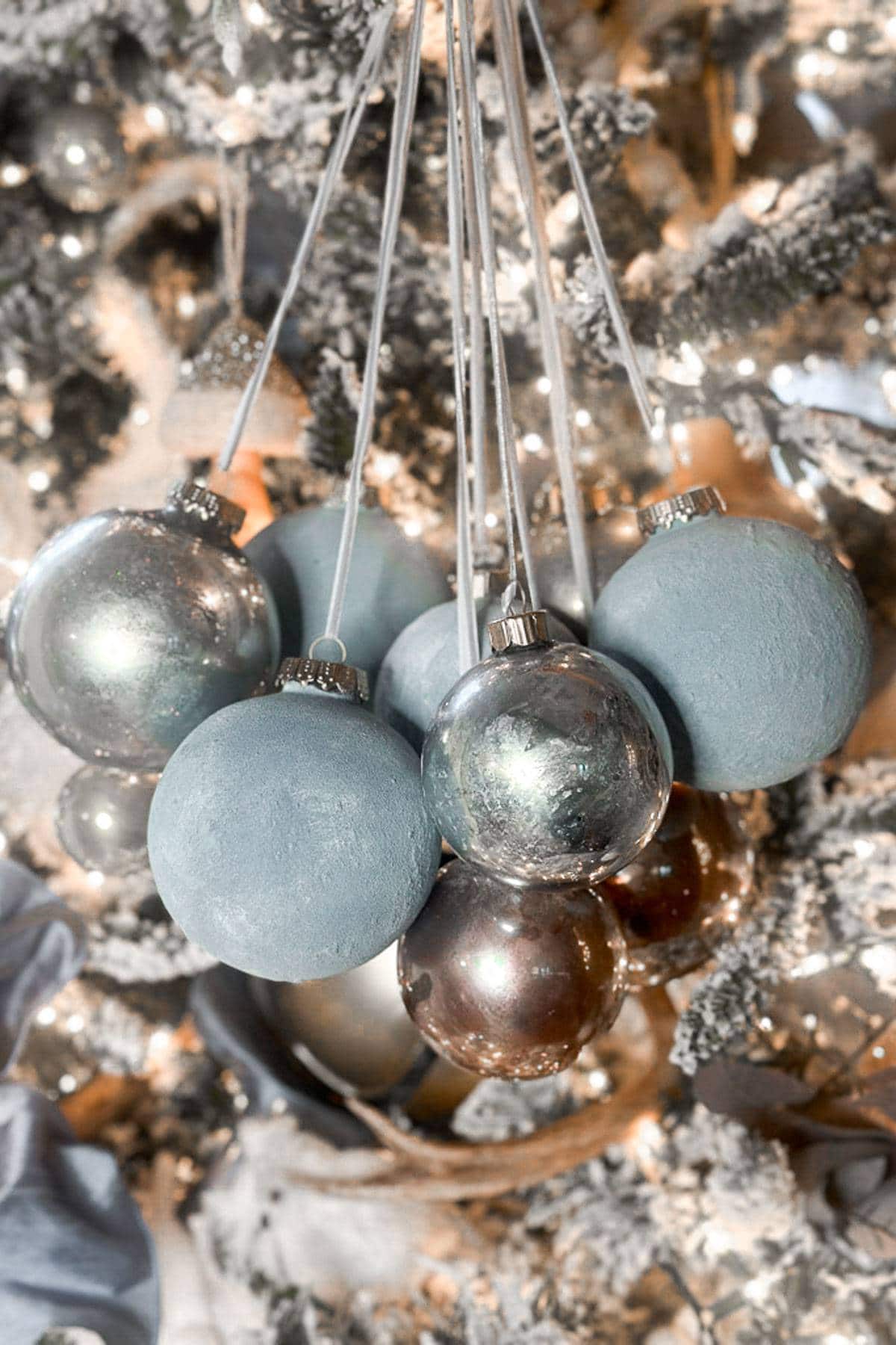 diy mercury glass ornaments-A cluster of blue and silver Christmas baubles hanging from strings in front of a decorated Christmas tree with twinkling lights. The background is softly blurred.