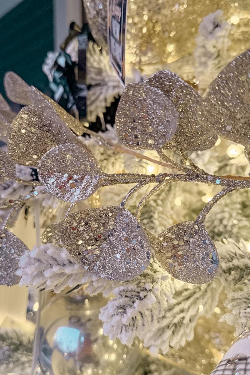 Close-up of a decorated Christmas tree featuring frosted branches and glittering leaves. The soft glow of warm white lights adds a festive sparkle to the scene, highlighting the tree's intricate details.