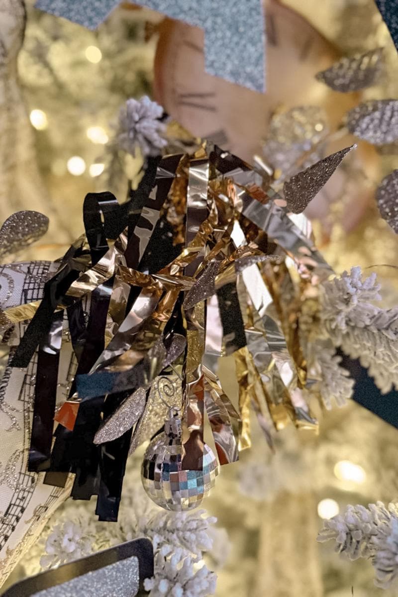 Close-up of a festive decoration featuring shiny metallic ribbons in silver and gold, a small disco ball ornament, and frosted pinecones, set against a softly lit background.