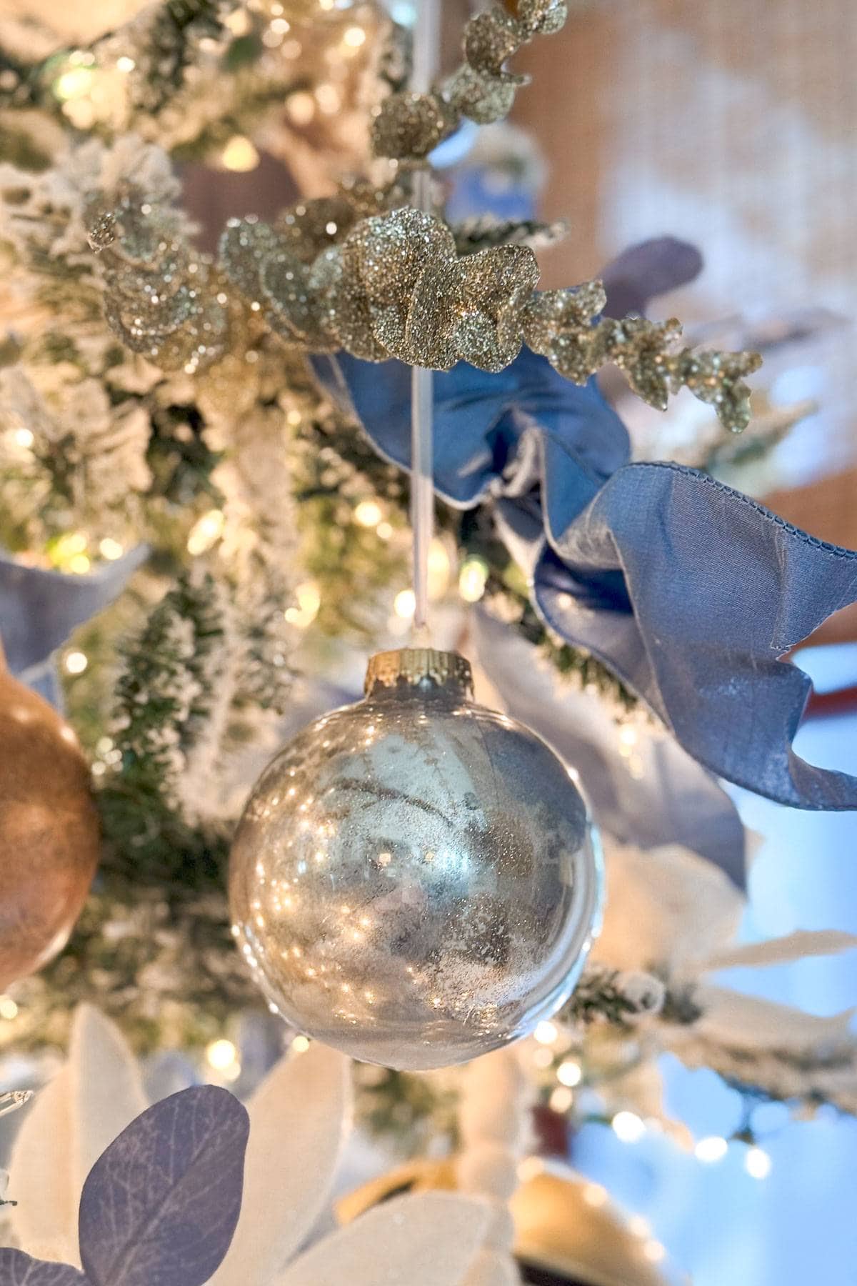 A close-up of a Christmas tree adorned with a shiny silver ornament. The tree is decorated with blue and gold ribbons, glittering foliage, and soft white lights, creating a festive and elegant holiday atmosphere.