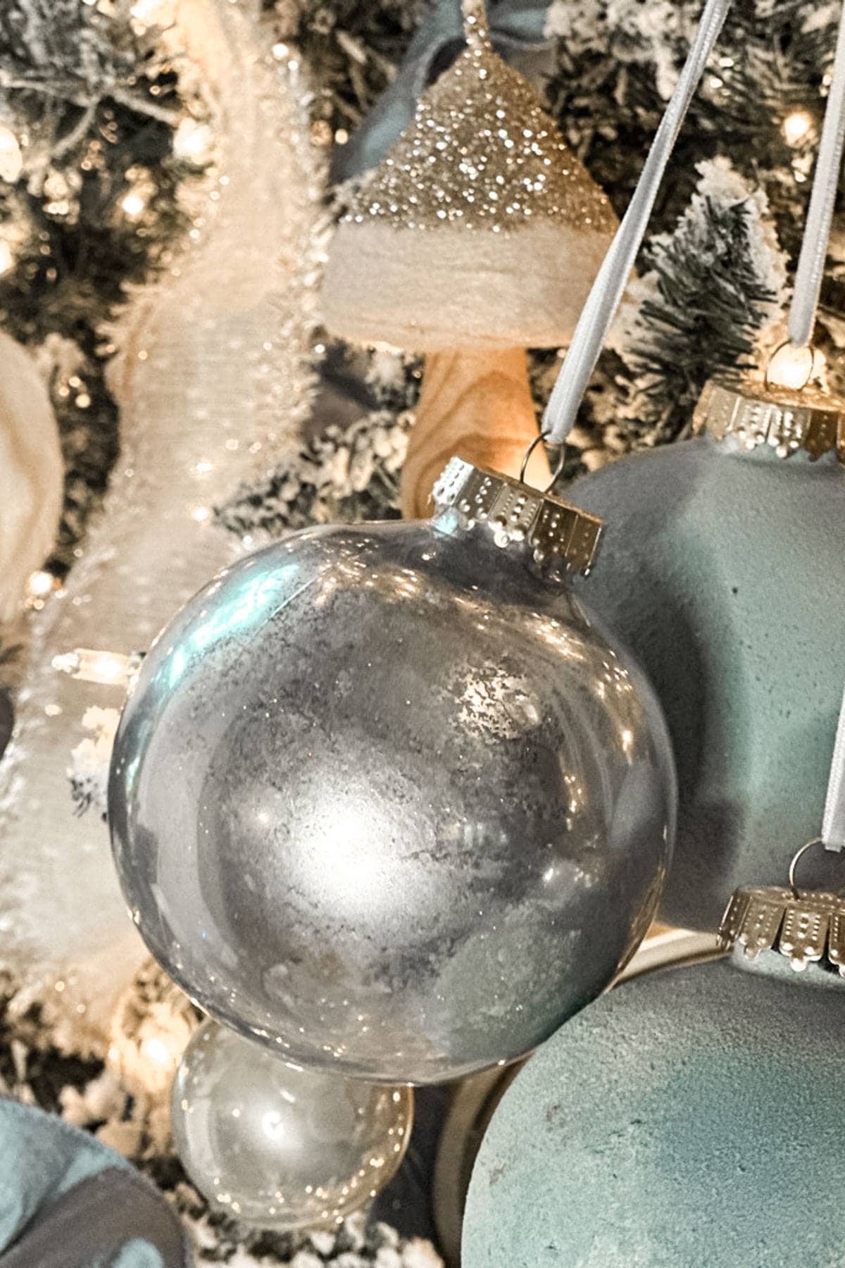 Close-up of a Christmas tree decorated with silver and teal ornaments. The ornaments have a shiny surface, reflecting light. The tree is adorned with white lights and other festive decorations, creating a bright, festive atmosphere.