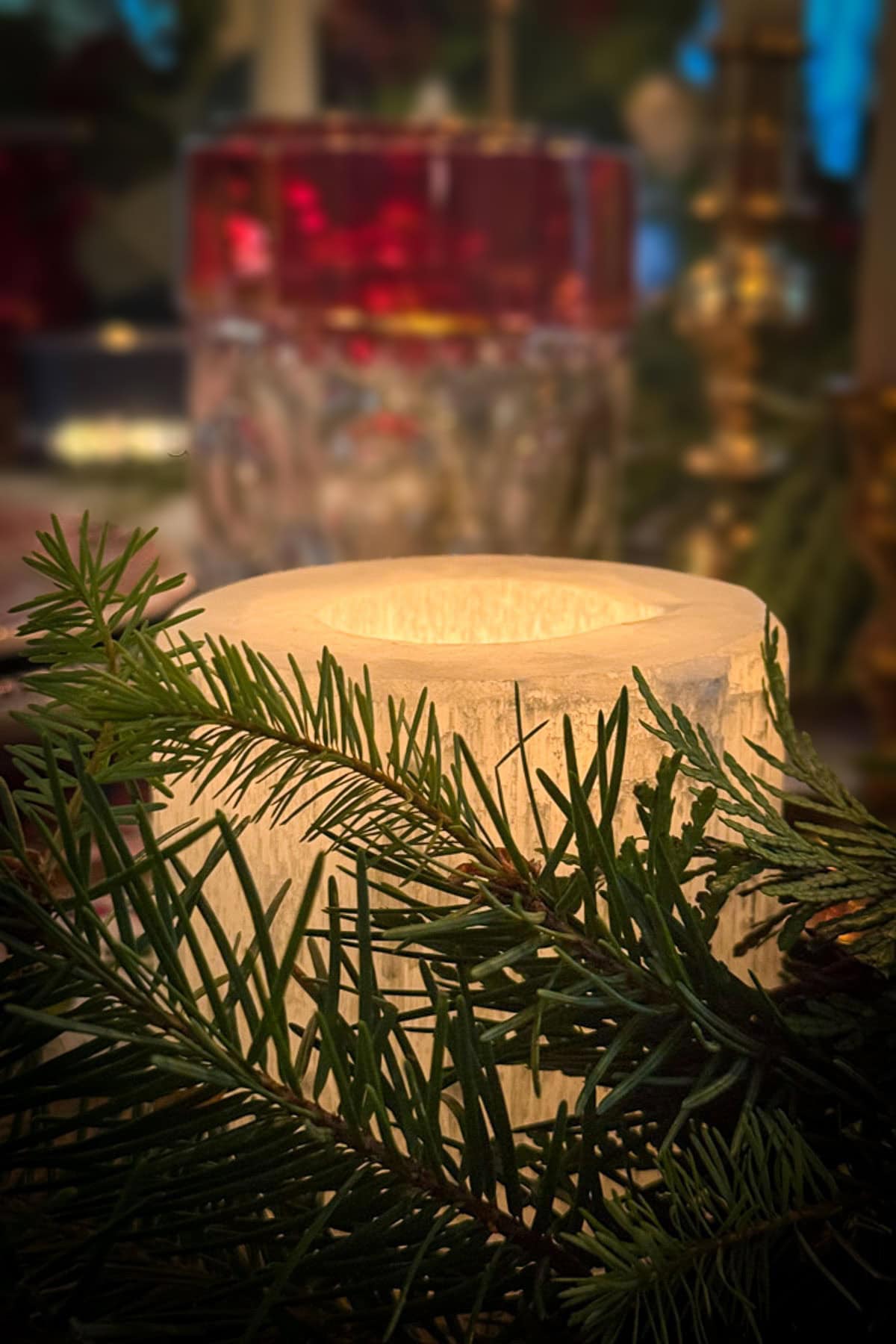 A lit white candle surrounded by pine branches in the foreground with a blurred red glass container and warm lighting in the background, creating a cozy, festive atmosphere.