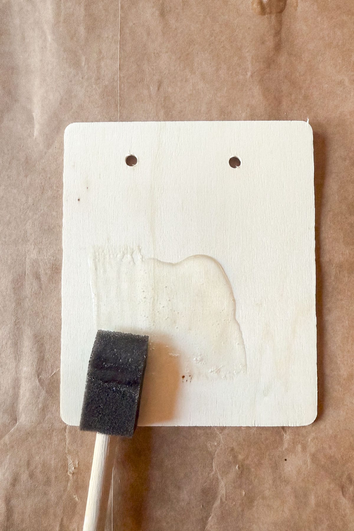 A wooden square with two holes near the top is positioned on a brown paper surface. A black foam brush with a wooden handle is applying a layer of clear varnish or paint to the wood, leaving a visible wet streak.