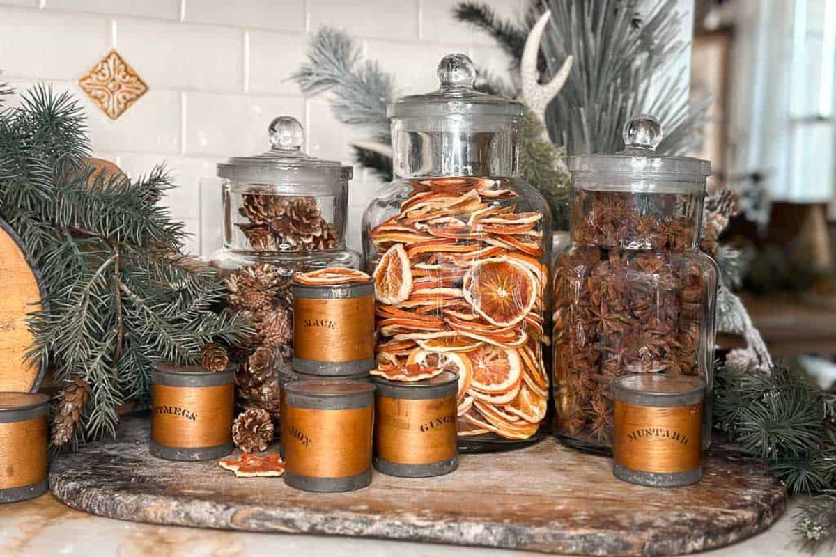 A rustic kitchen display featuring glass jars filled with dried oranges, cinnamon sticks, and pinecones, surrounded by evergreen branches. Several small brown spice jars are arranged on a wooden board.