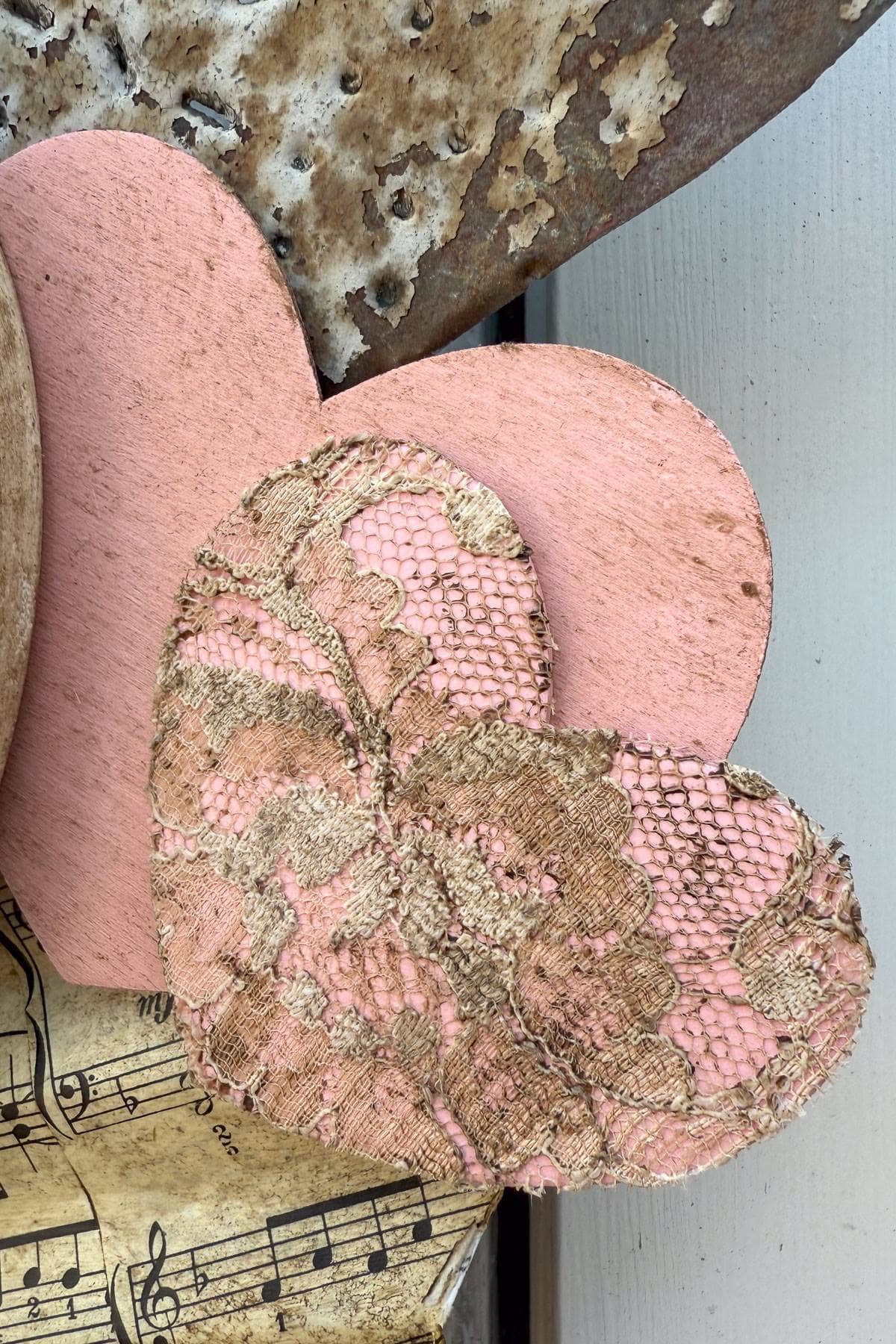 Two heart-shaped objects, one painted pink and the other covered with pink and gold lace, rest on a weathered surface. Below, there is a portion of sheet music, adding a vintage feel to the composition.