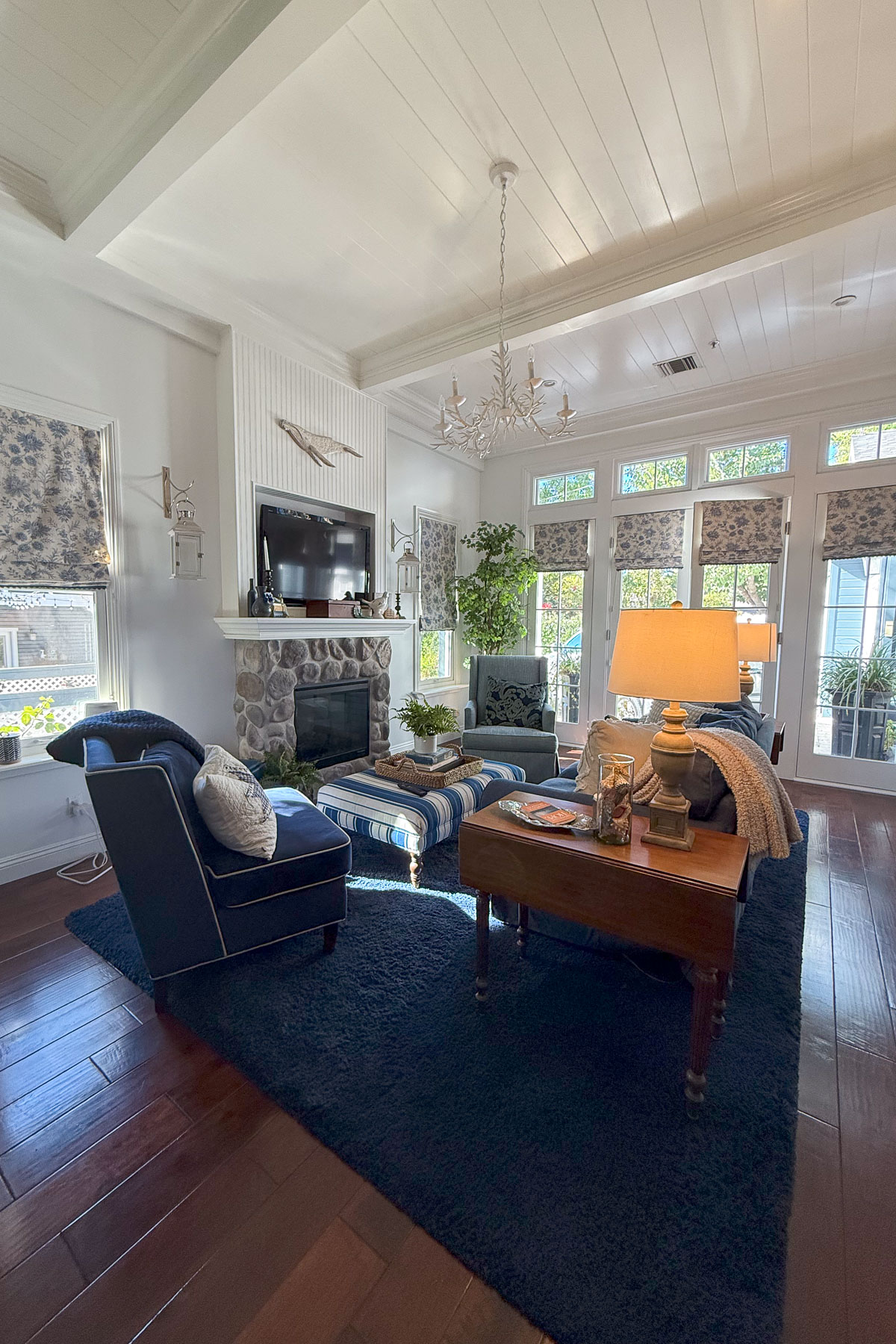 A cozy living room with a navy blue sectional sofa, striped ottoman, and coffee table on a dark carpet. A stone fireplace with a mounted TV is in the background, framed by windows with floral drapes. A chandelier hangs from the ceiling.