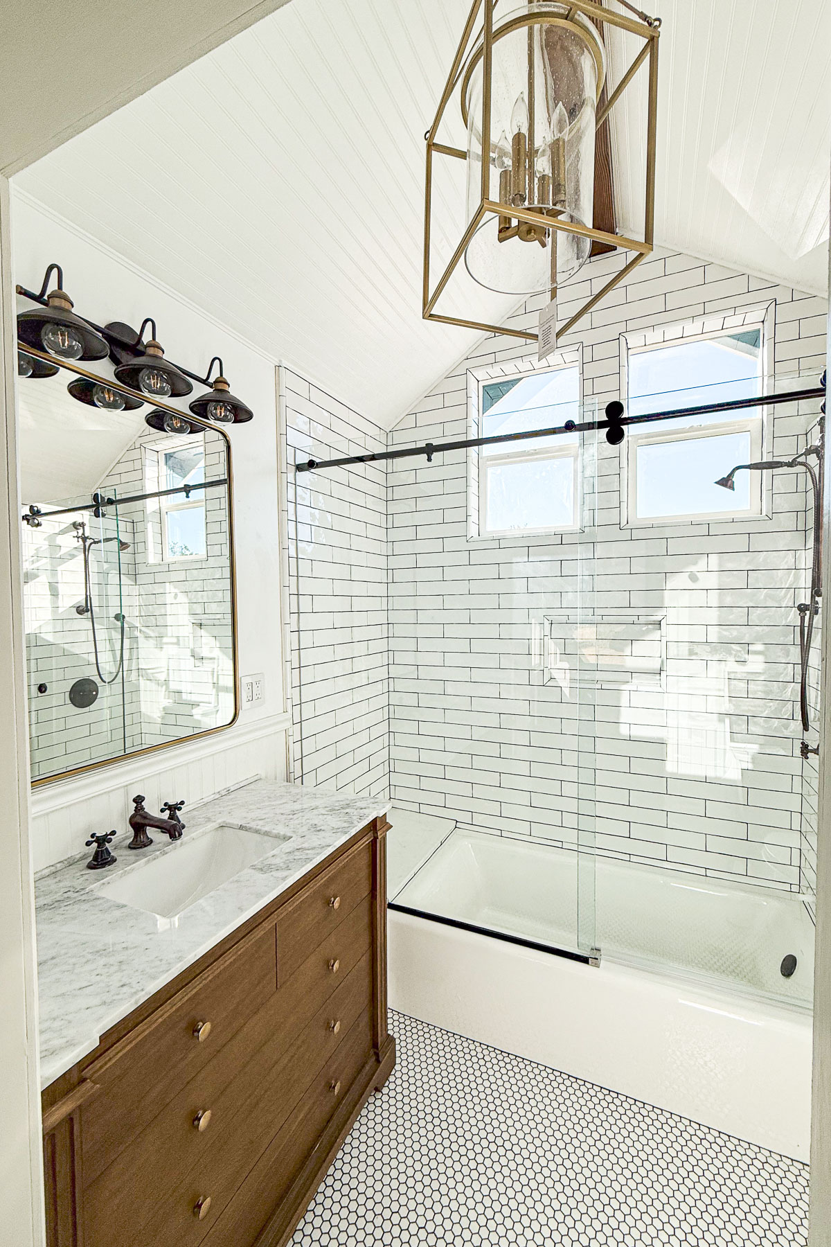 A bright bathroom featuring white subway tiles, a glass-enclosed bathtub, a wooden vanity with a marble countertop, and a hexagonal tile floor. The space is illuminated by a modern chandelier and natural light from two small windows.