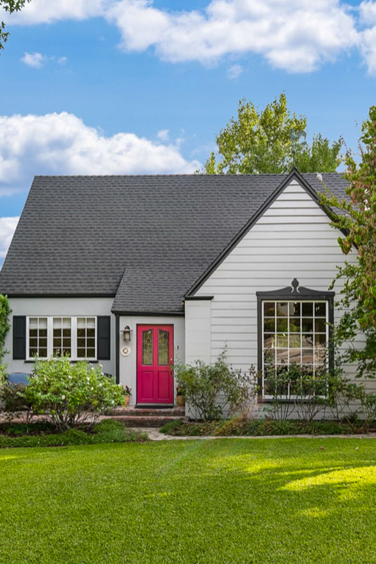 A charming white house with a dark gray gabled roof and vibrant red front door. It features black shutters on the left window and has a well-maintained green lawn with shrubs. The sky is partly cloudy, adding to the picturesque setting.