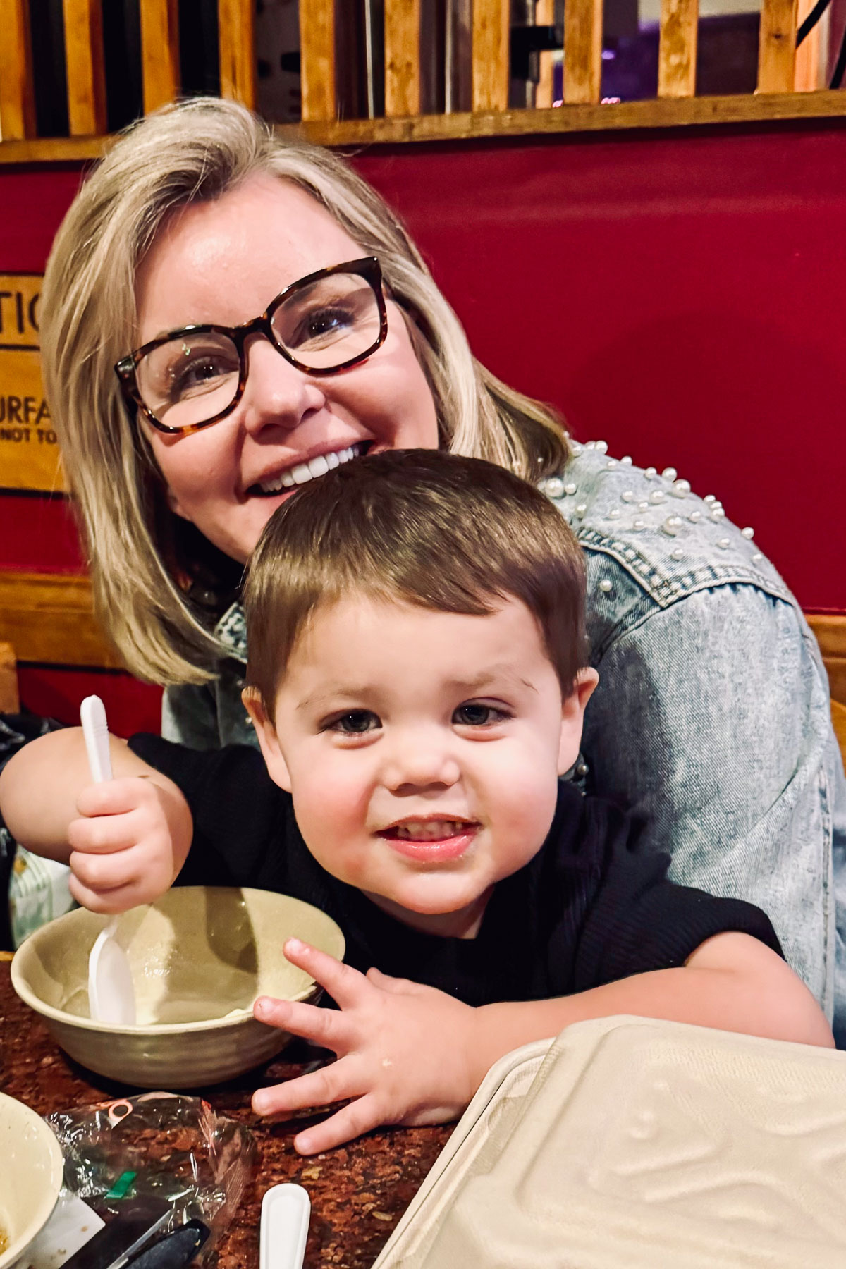 A woman with glasses and a young child are smiling at a table in a restaurant. The child is holding a spoon over a bowl. They are sitting close together, and there is a wooden railing in the background.