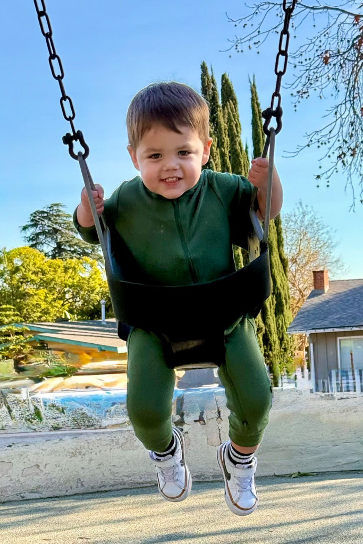 A toddler with short brown hair and a green outfit smiles while swinging in a playground swing, enjoying the day as if it were Sunday Sharing. The background includes trees, a house resembling a cozy she shed, and a clear blue sky.