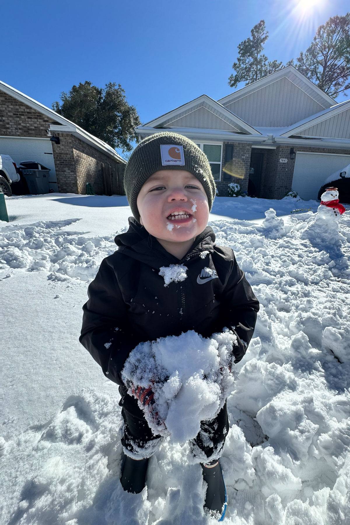 A toddler in a green beanie and black jacket joyfully holds a snowball in a snowy front yard. Sunlight shines on their smiling face. Houses and a snowman are visible in the background under a clear blue sky.