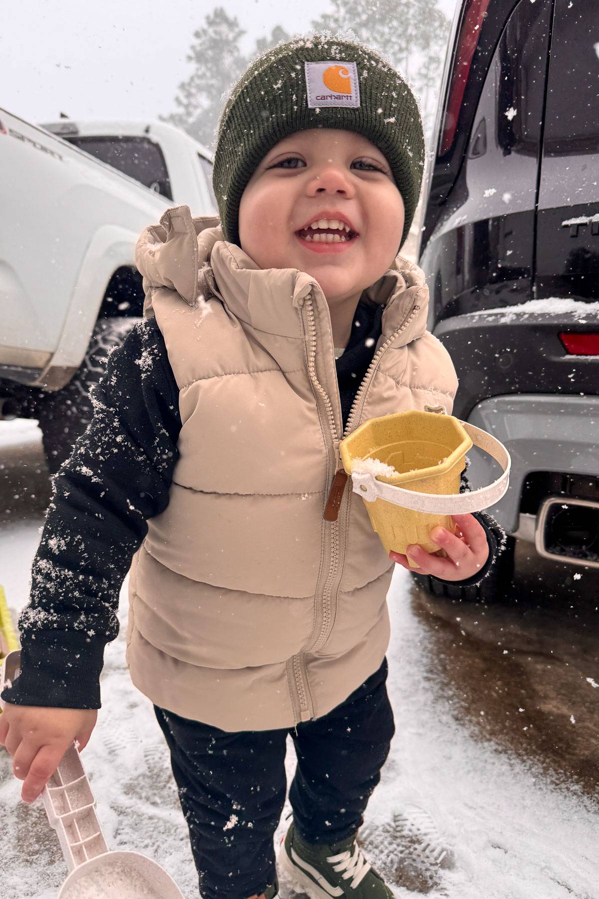 Child smiling in the snow, wearing a beige puffy vest, dark pants, and a green beanie. They're holding a toy bucket and surrounded by parked cars. Snowflakes are falling gently.