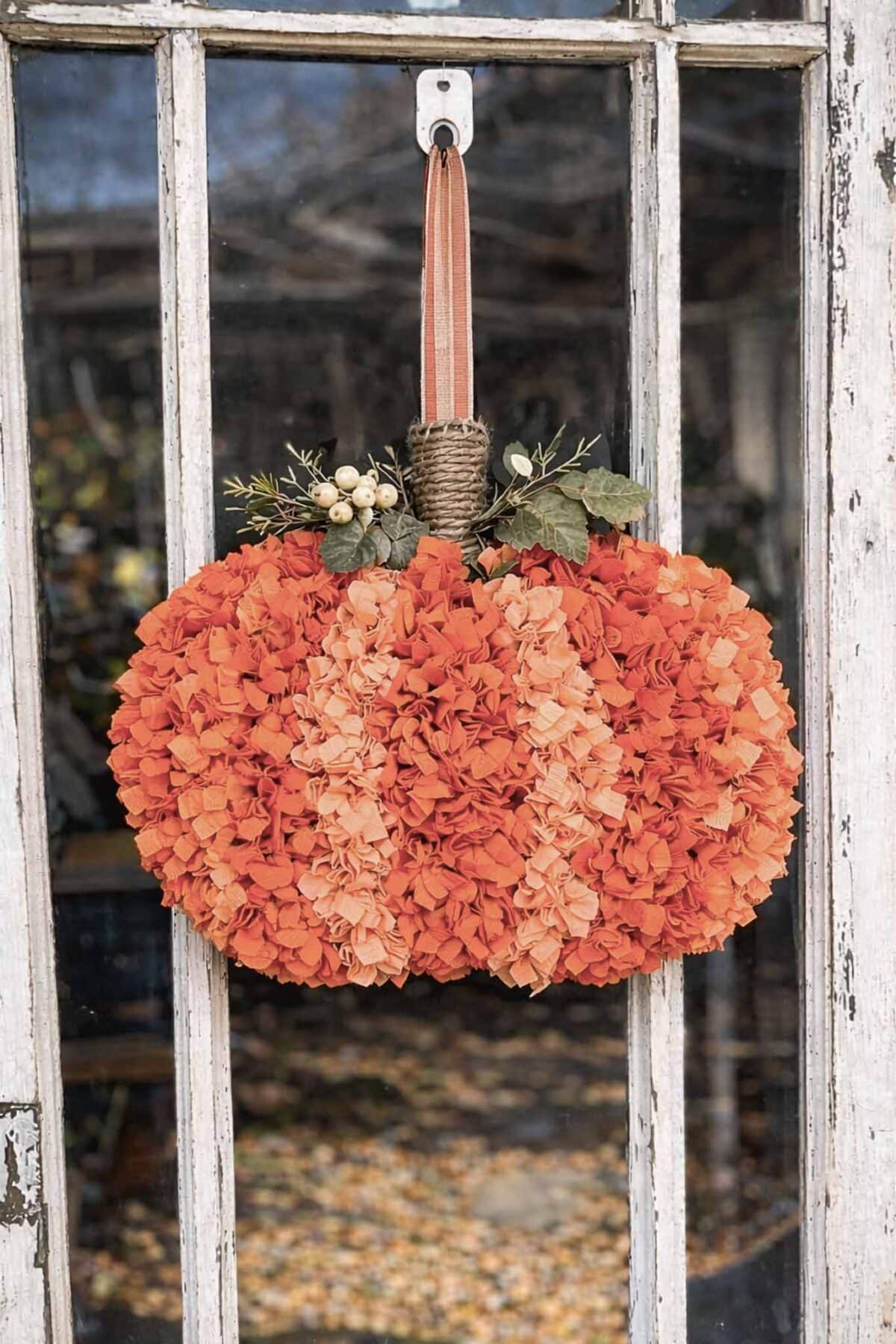 A decorative DIY Rag Wreath shaped like a pumpkin, made of orange and peach flowers, hangs on a window by a brown ribbon. Green leaves and white berries are attached at the top for added detail.