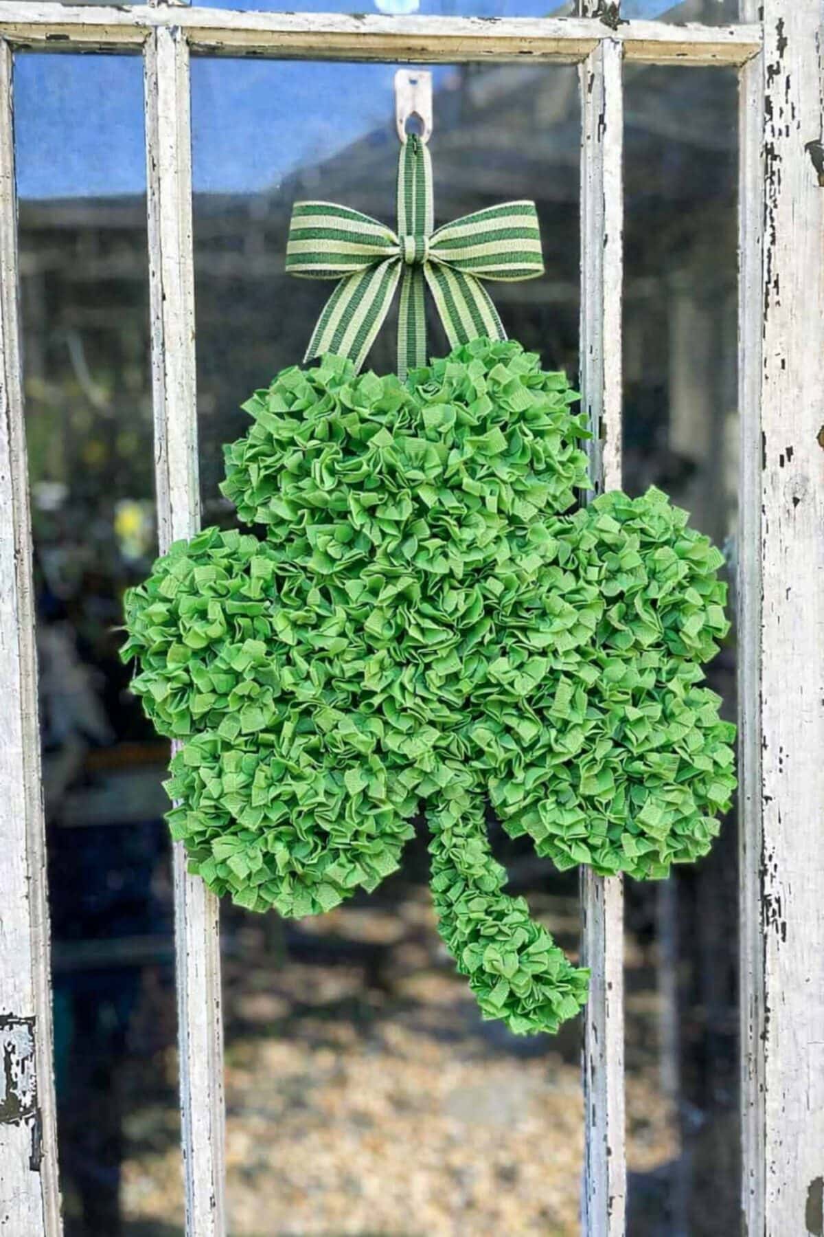 A decorative DIY Rag Wreath made of green fabric pieces hangs on a weathered glass door. The wreath, shaped like a three-leaf clover, features a striped green bow at the top for a festive shamrock touch.