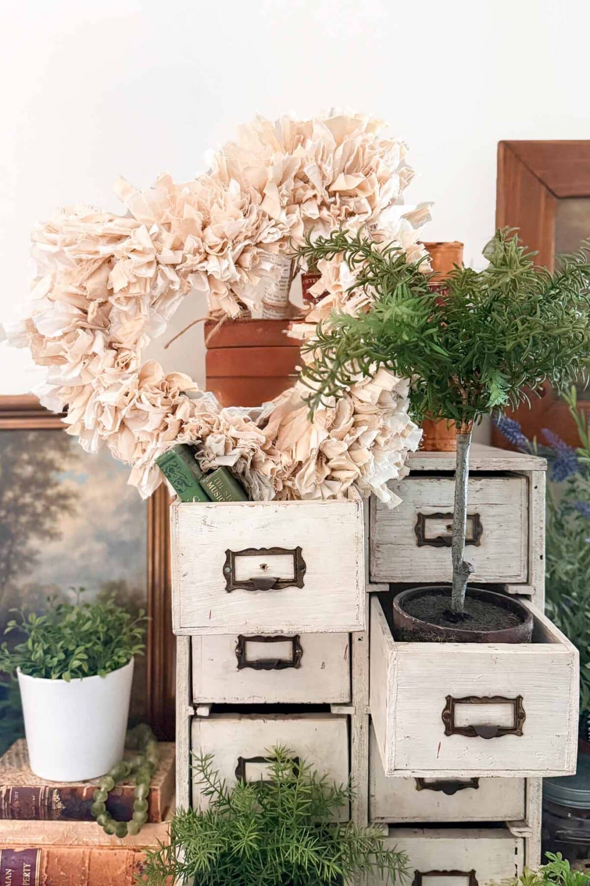 A white vintage drawer unit with potted plants and a beige DIY Rag Wreath heart on top. One drawer is open, holding a small tree in a pot. Books and additional plants are arranged nearby.
