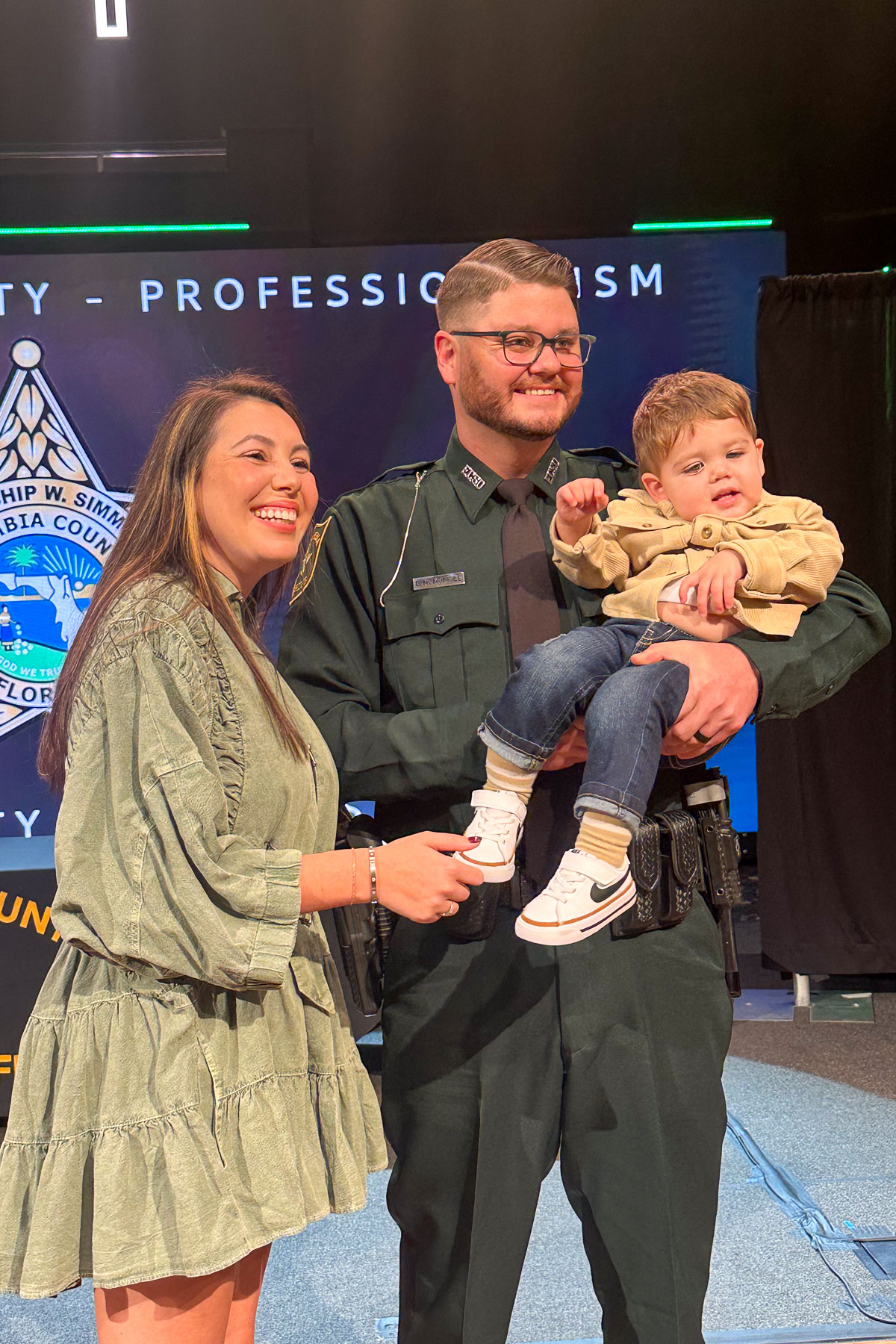 A smiling woman stands next to a man in a dark green uniform holding a young child. They appear happy and are in front of a backdrop with text and a logo.