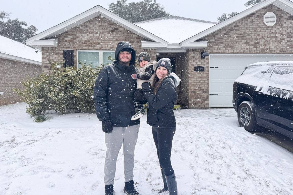 A family of three stands outside a brick house on a snowy day. They are dressed warmly in winter clothing. Snow is falling, and the ground is covered in a layer of snow. The house and car in the background are partially dusted with snow.