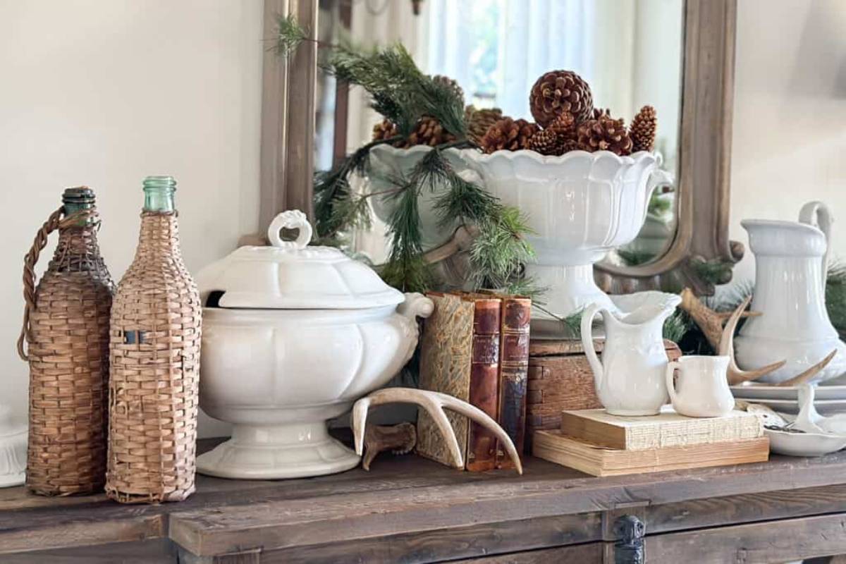 A rustic decor arrangement on a wooden shelf with woven bottles, white ceramic dishes, pinecones, evergreen branches, vintage books, and antlers. A mirror reflects the display, adding depth to the cozy, vintage-inspired scene.