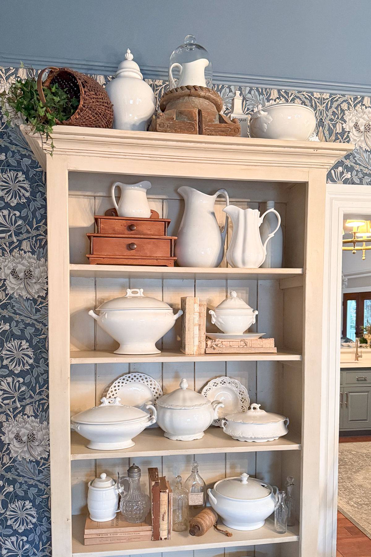 A vintage cupboard displays white ceramic pitchers, platters, and tureens. Wooden boxes and old books are also arranged on the shelves. The background features blue and white floral wallpaper, and a wicker basket with greenery sits on top.