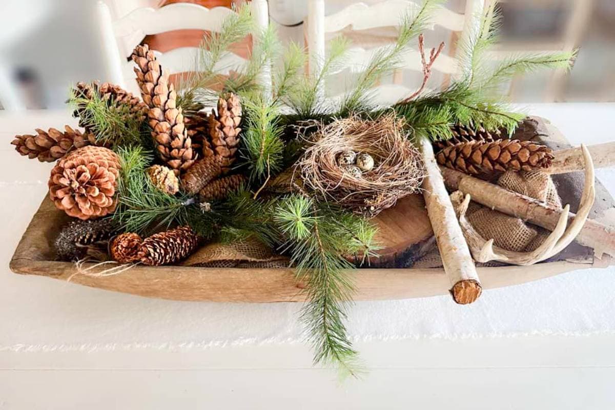 A rustic wooden tray centerpiece with pine cones, green pine branches, a bird's nest with small eggs, and decorative antlers. There's also a small bundle of birch sticks, creating a natural, woodland-themed display.