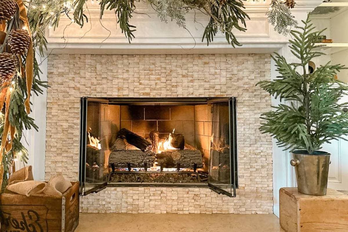 A cozy fireplace with a lit fire is surrounded by a stone mantel. Pinecone and evergreen garlands adorn the mantel, and a small potted evergreen tree stands nearby. A wooden box with logs is placed beside the fireplace.