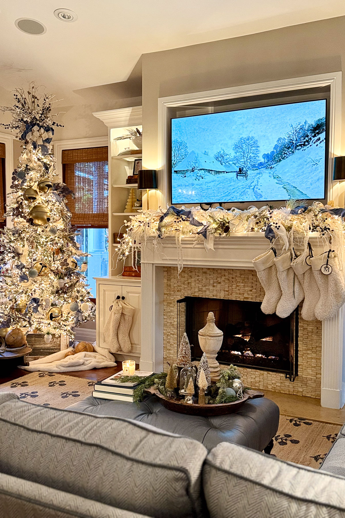 A cozy living room decorated for Christmas features a lit tree adorned with white and gold ornaments, evoking the warmth of a Sunday Sharing. Stockings hang over the fireplace, with a TV displaying a snowy scene. Decorative trees dot the coffee table, while soft lighting enhances the inviting ambiance.