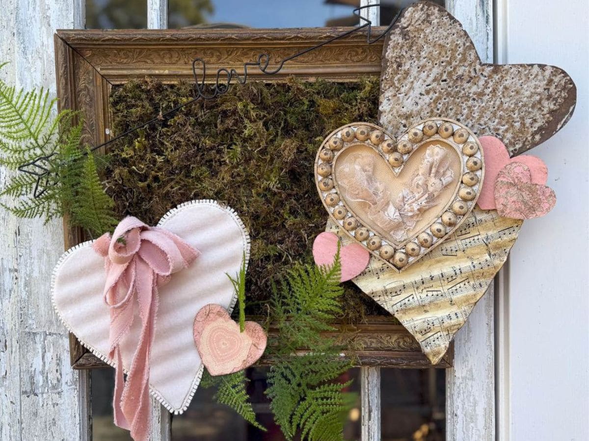 A decorative collage inspired by Valentine decor features heart shapes with various textures: pink fabric, musical notes, and rustic metal. Fern leaves and a mossy background frame the arrangement, with wire spelling "love" at the top. A unique vintage touch enhances this DIY decorating idea.