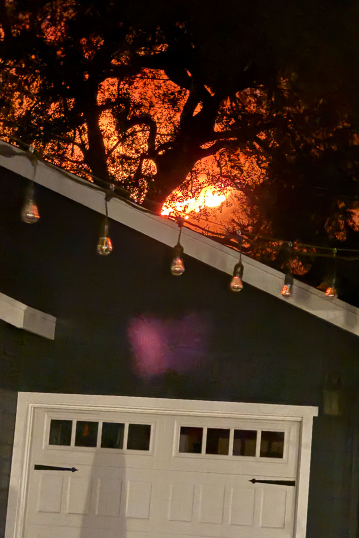 A garage with a white door is in the foreground, adorned with hanging string lights. Behind it, a large fire is visible through silhouetted trees, casting an orange glow into the night sky.