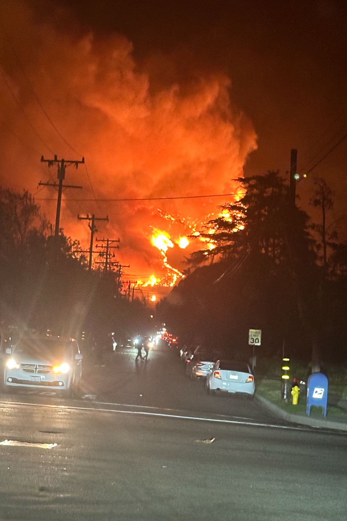 A large fire with intense orange flames and thick smoke billows into the night sky. The fire is visible down a residential street lined with parked cars. Utility poles and silhouetted trees are in the foreground, with a few people walking on the street.