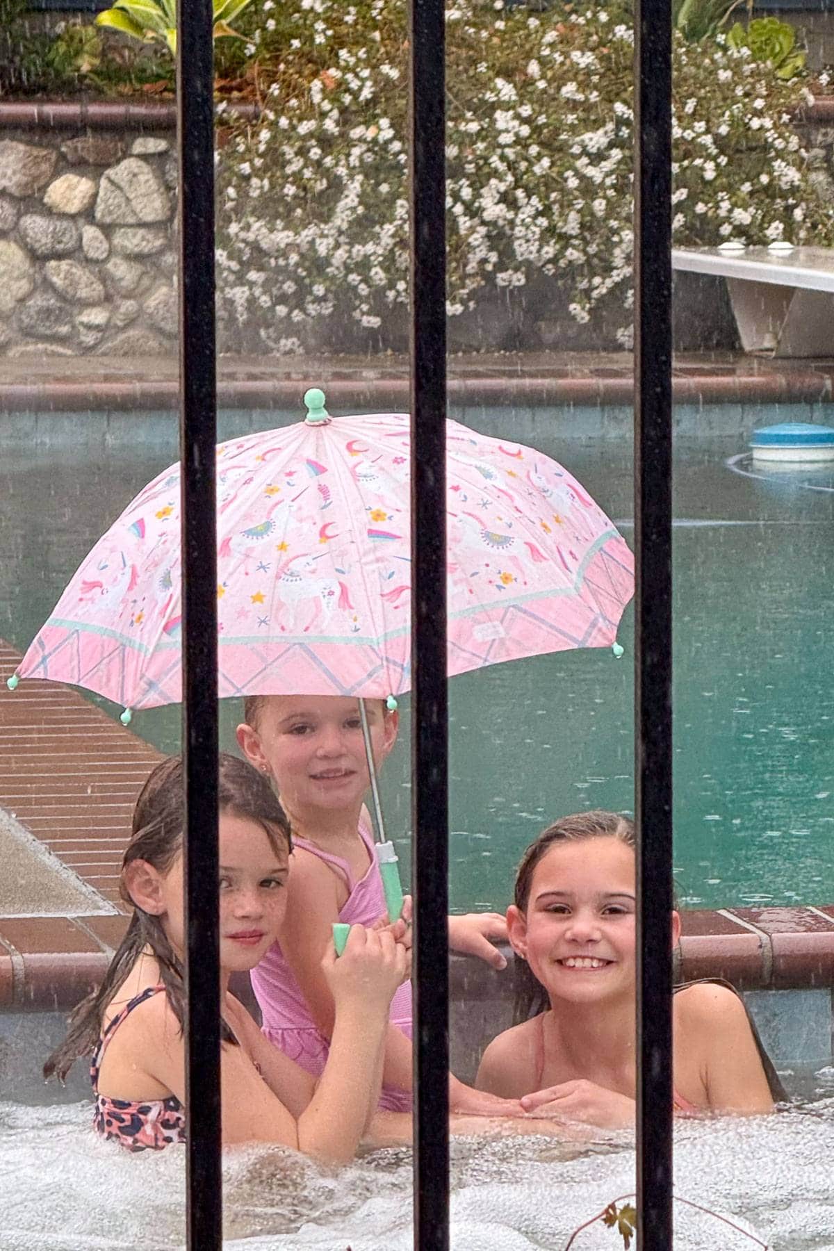 Three children sit in a hot tub under a pink umbrella, smiling at the camera. They are surrounded by rain, with a swimming pool and stone wall in the background. Black railings frame the foreground.