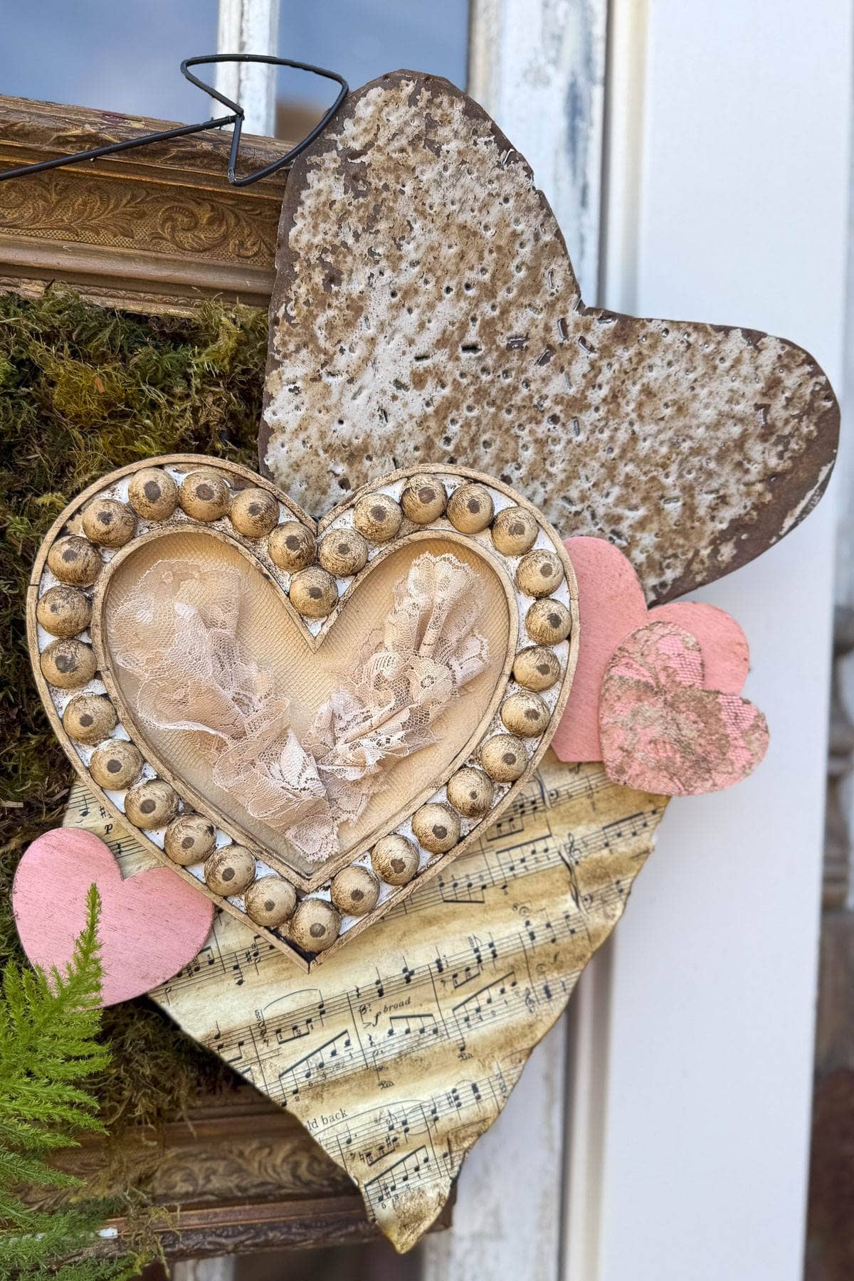 A decorative arrangement featuring layered hearts: a large rustic heart, an ornate heart with lace and pearls, smaller pink hearts, and a heart made from sheet music. Set against a backdrop with moss and a partial frame.