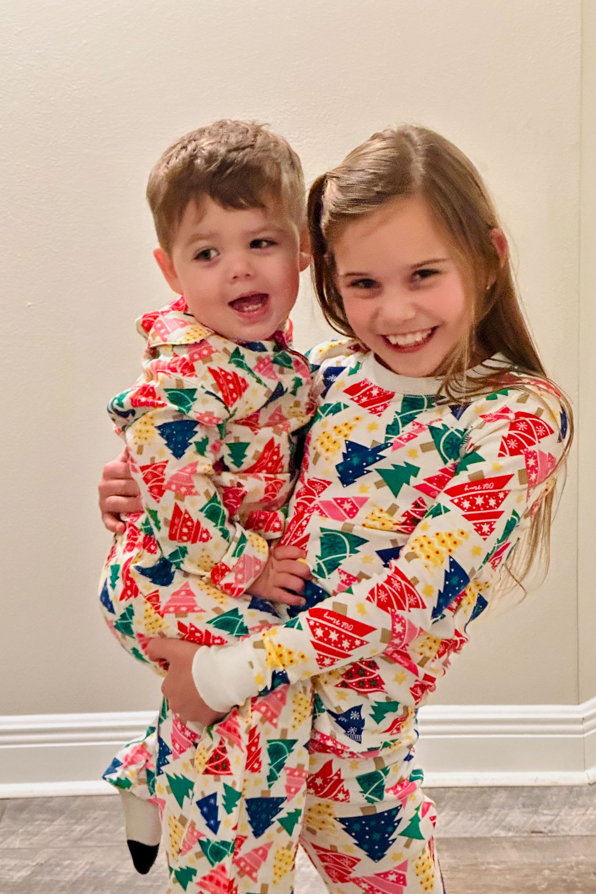 A young girl is joyfully holding a toddler. Both are wearing matching festive pajamas with colorful trees and shapes. They are standing indoors on a wooden floor in front of a plain wall.