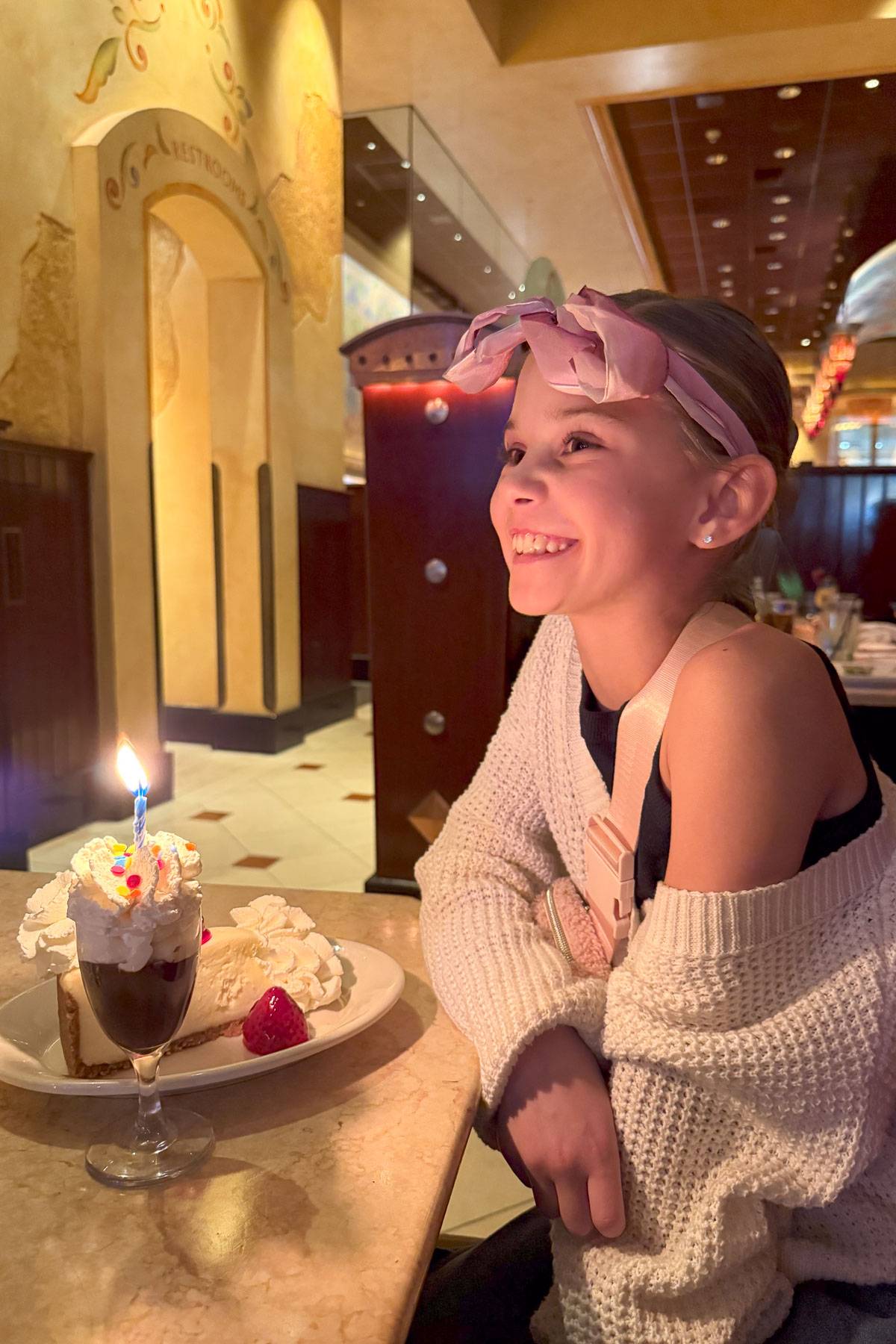 A smiling girl wearing a pink headband sits at a restaurant table, wearing a white sweater. In front of her is a dessert with a lit candle and a slice of cake topped with whipped cream and a cherry. The restaurant interior is warmly lit.