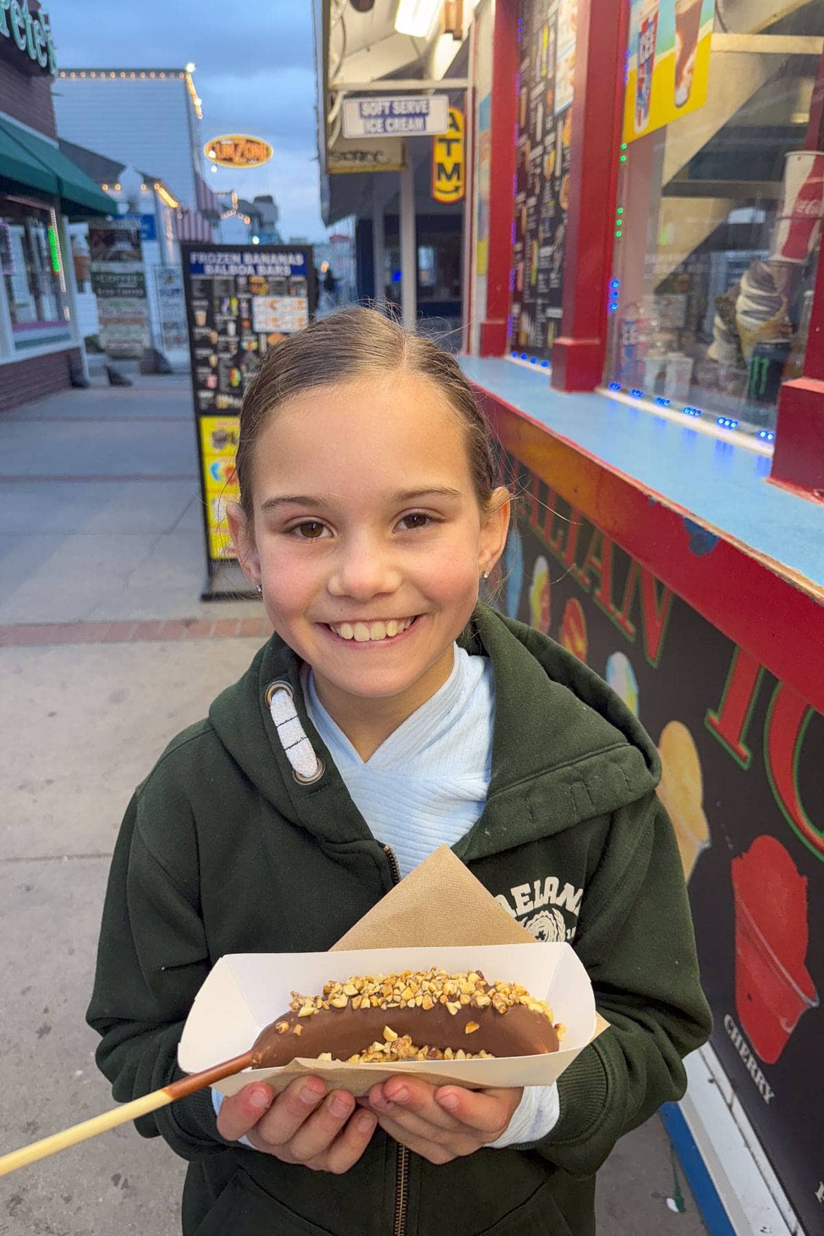 A smiling child in a green hoodie holds a chocolate-covered treat on a stick, topped with nuts. They stand in front of a colorful food stand on a sidewalk, with shops visible in the background.