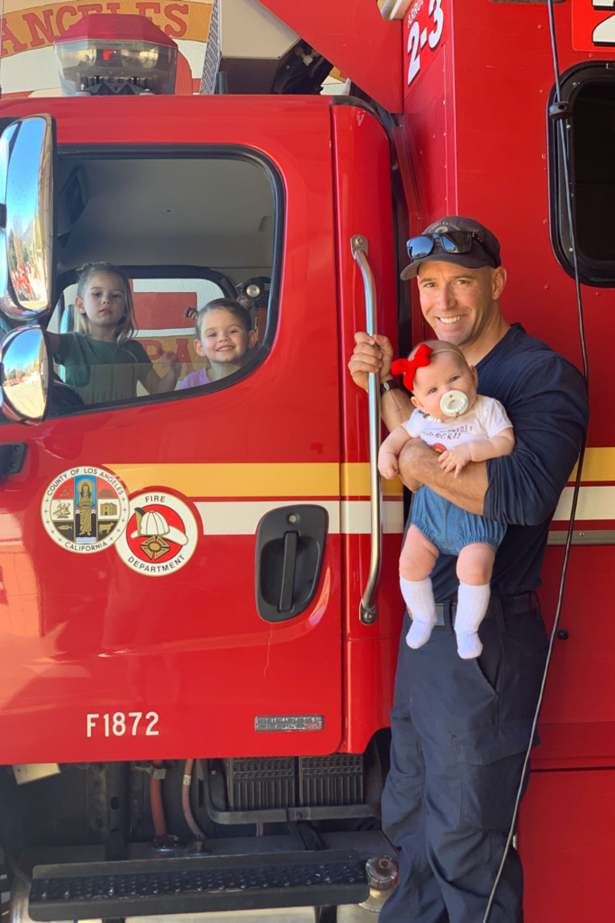 A smiling firefighter stands beside a red fire truck holding a baby in his arms. Two young children are sitting inside the truck, looking out of the window. The fire department emblem is visible on the truck's door.