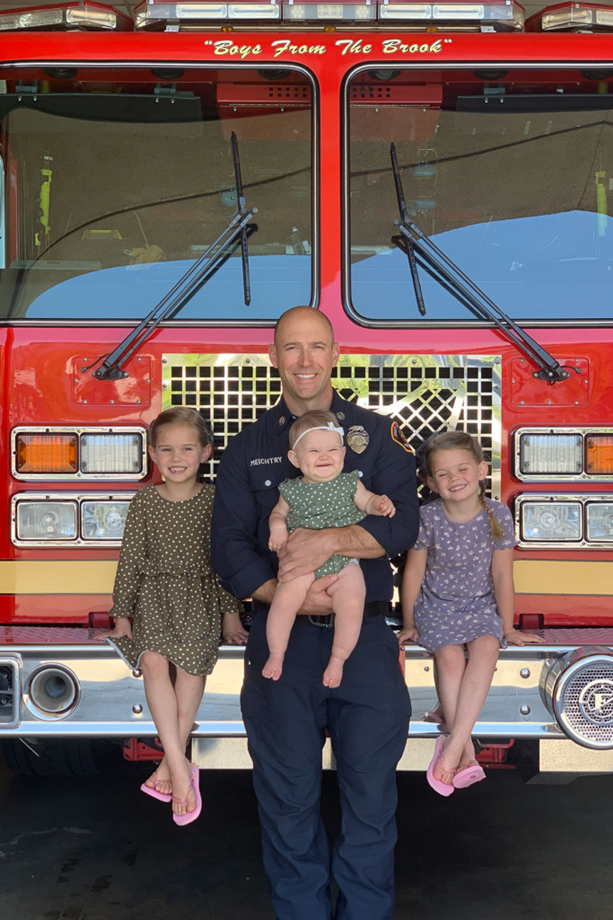 A smiling firefighter stands in front of a bright red fire truck with three children. Two young girls in polka dot dresses sit on either side of him, and he holds a baby in a light outfit. The words "Boys From The Bronx" are visible on the truck.