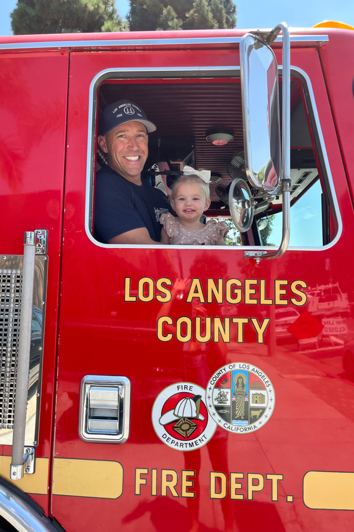 A smiling firefighter and a young child sit inside a bright red Los Angeles County Fire Department truck. The firefighter is wearing a blue uniform and cap, while the child is wearing a pink outfit. The truck door displays the fire department's logo.