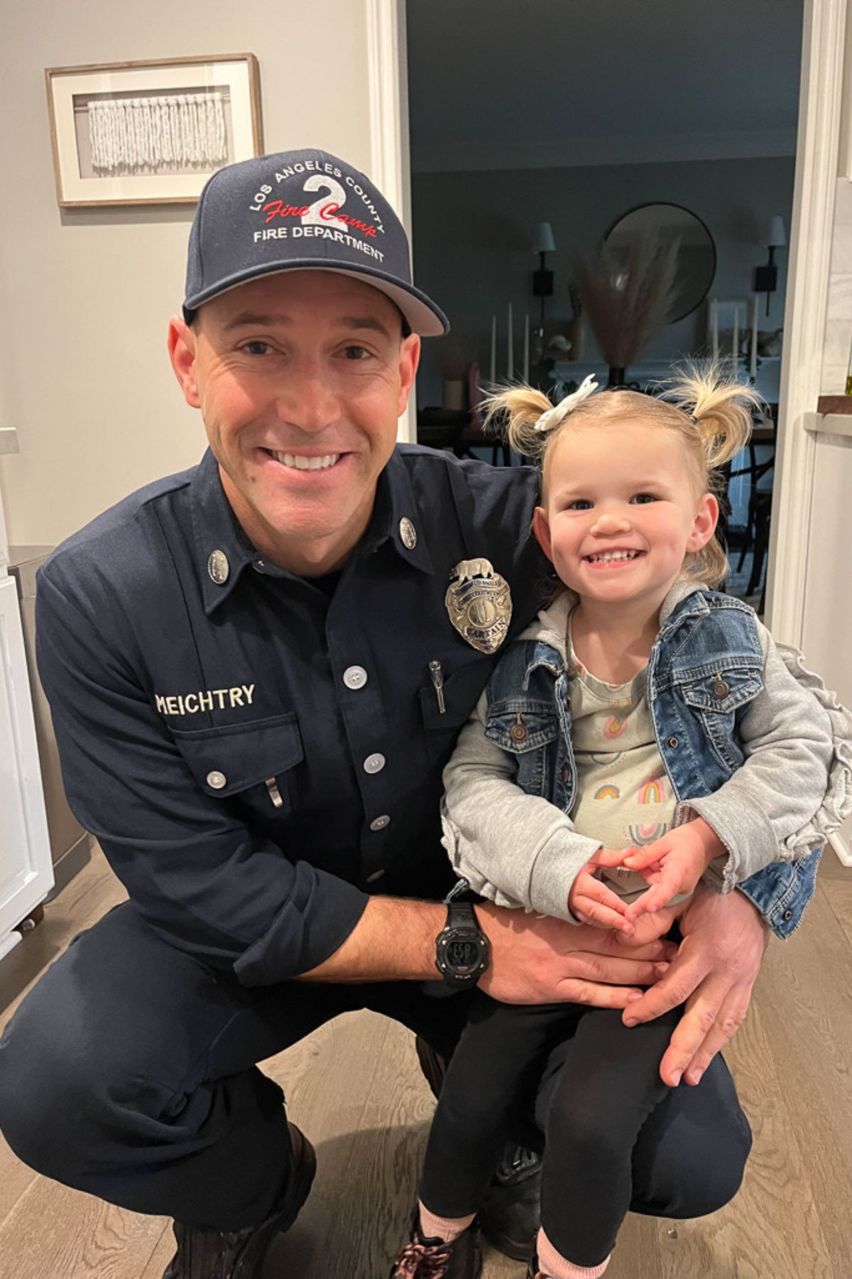 A firefighter in uniform kneels and smiles next to a young girl with pigtails and a denim jacket. They are indoors, with a framed picture and a dining area in the background.