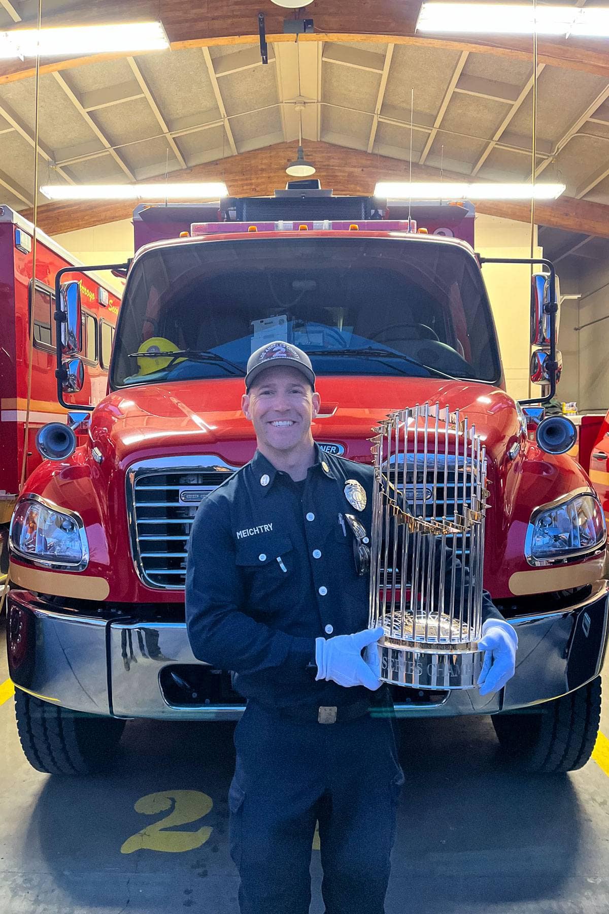 A firefighter in a navy uniform and white gloves holds a large trophy in front of a red fire truck inside a garage. The garage has a high, wooden ceiling and bright lights.