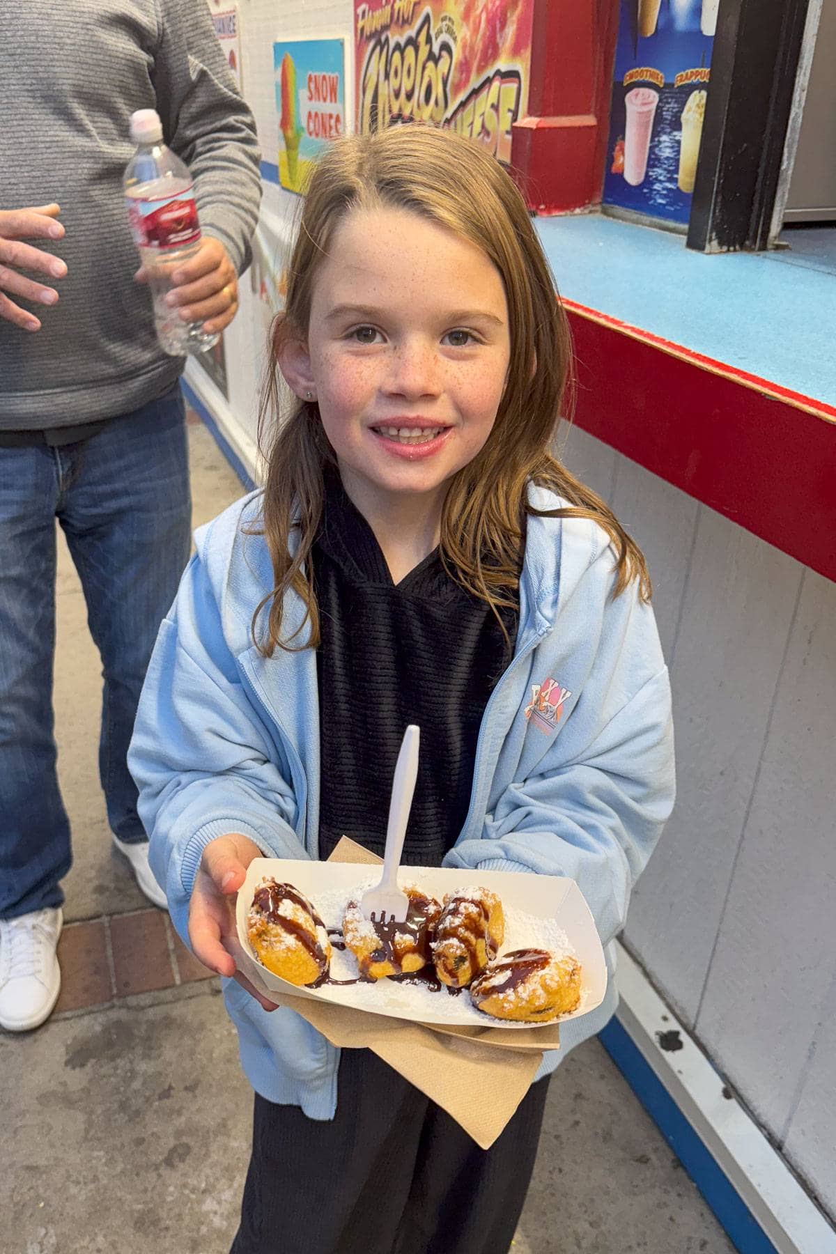 A young girl stands smiling, holding a tray of food with a fork. The tray contains several fried snacks drizzled with chocolate sauce. She is wearing a light blue jacket. There is a person partly visible on her right holding a water bottle.