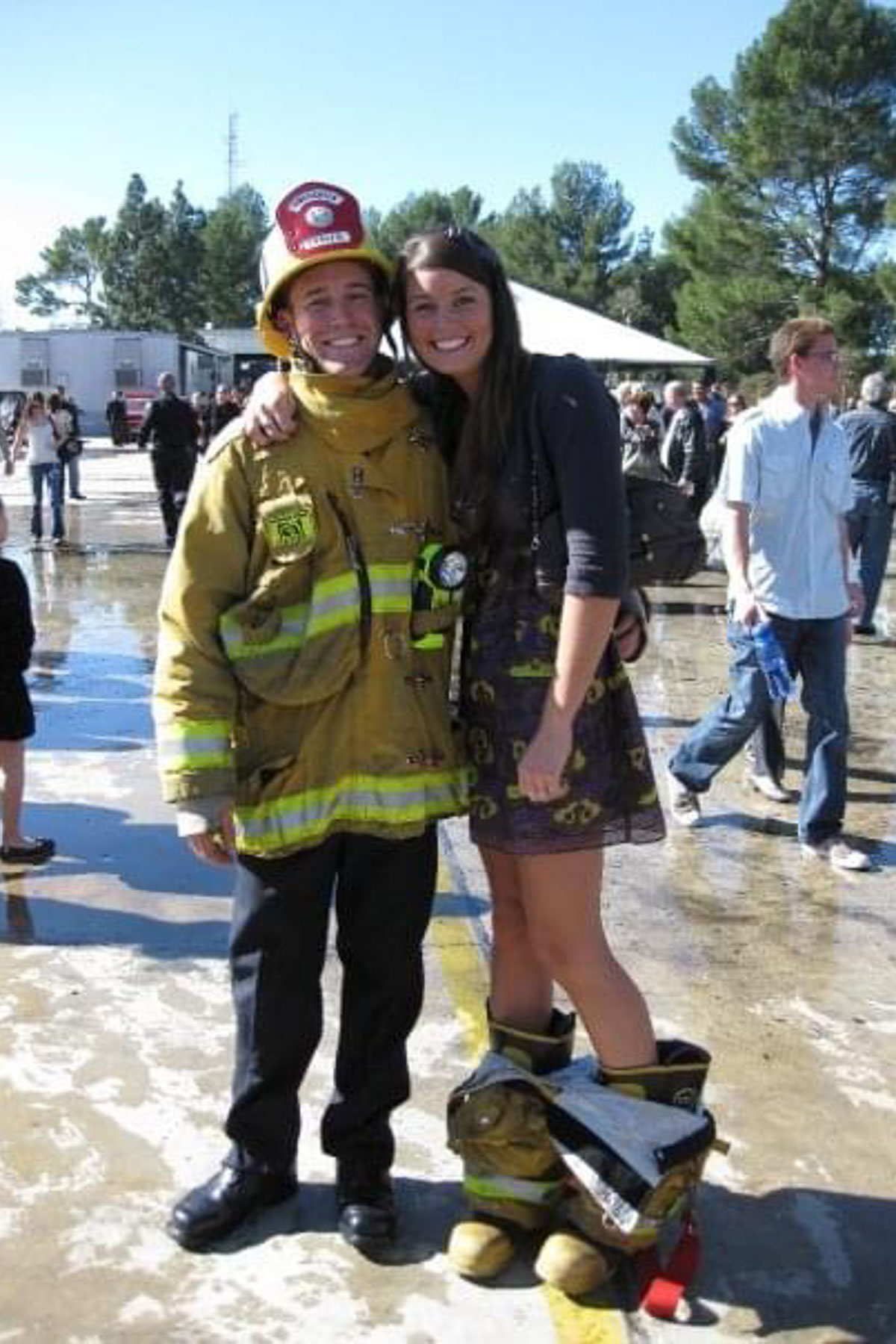 A firefighter in full gear stands smiling beside a woman wearing oversized firefighter pants and boots, clinging to her partner's arm. They are outdoors on a wet surface with trees and people in the background.