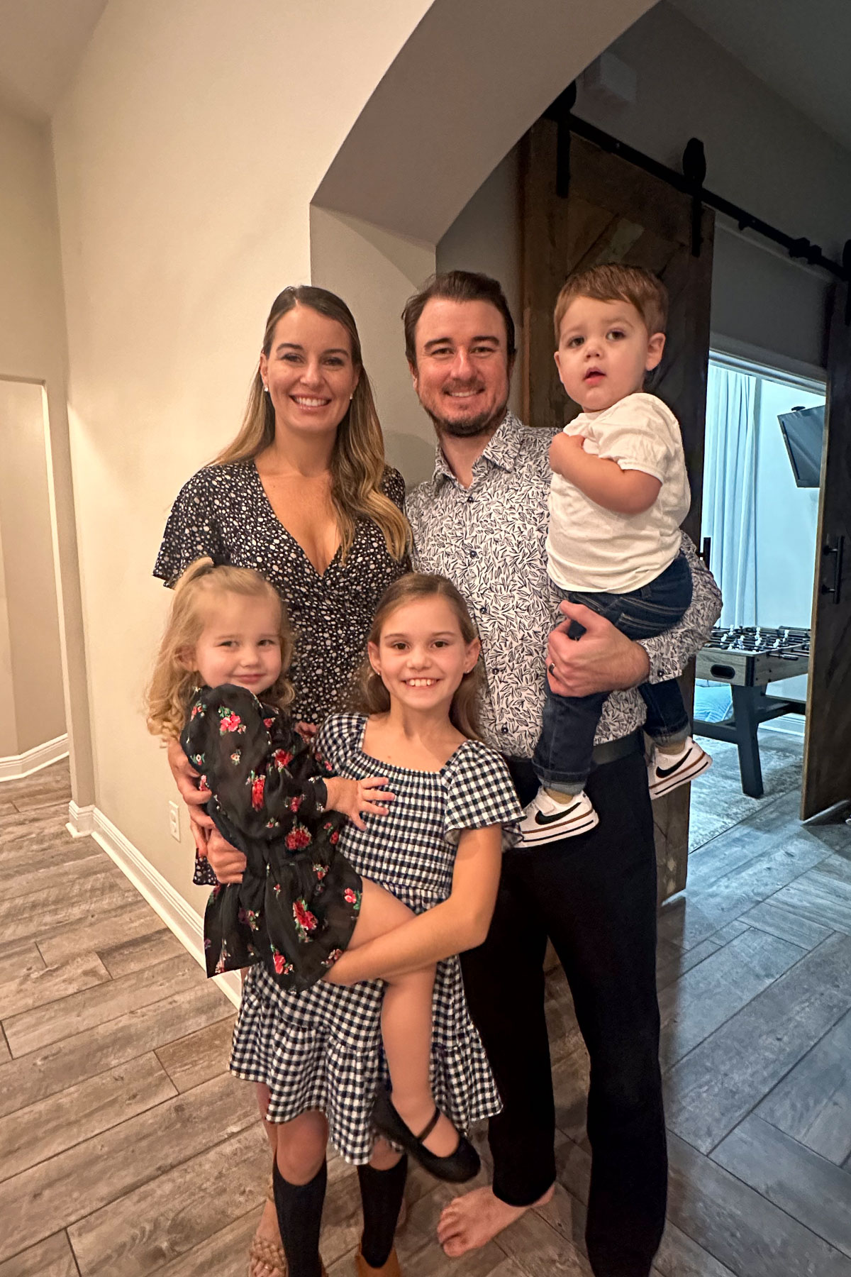 A smiling family stands indoors. The mother holds a young girl wearing a floral dress. The father holds a toddler boy and stands next to another girl in a checkered dress. They are all casually dressed and appear happy.