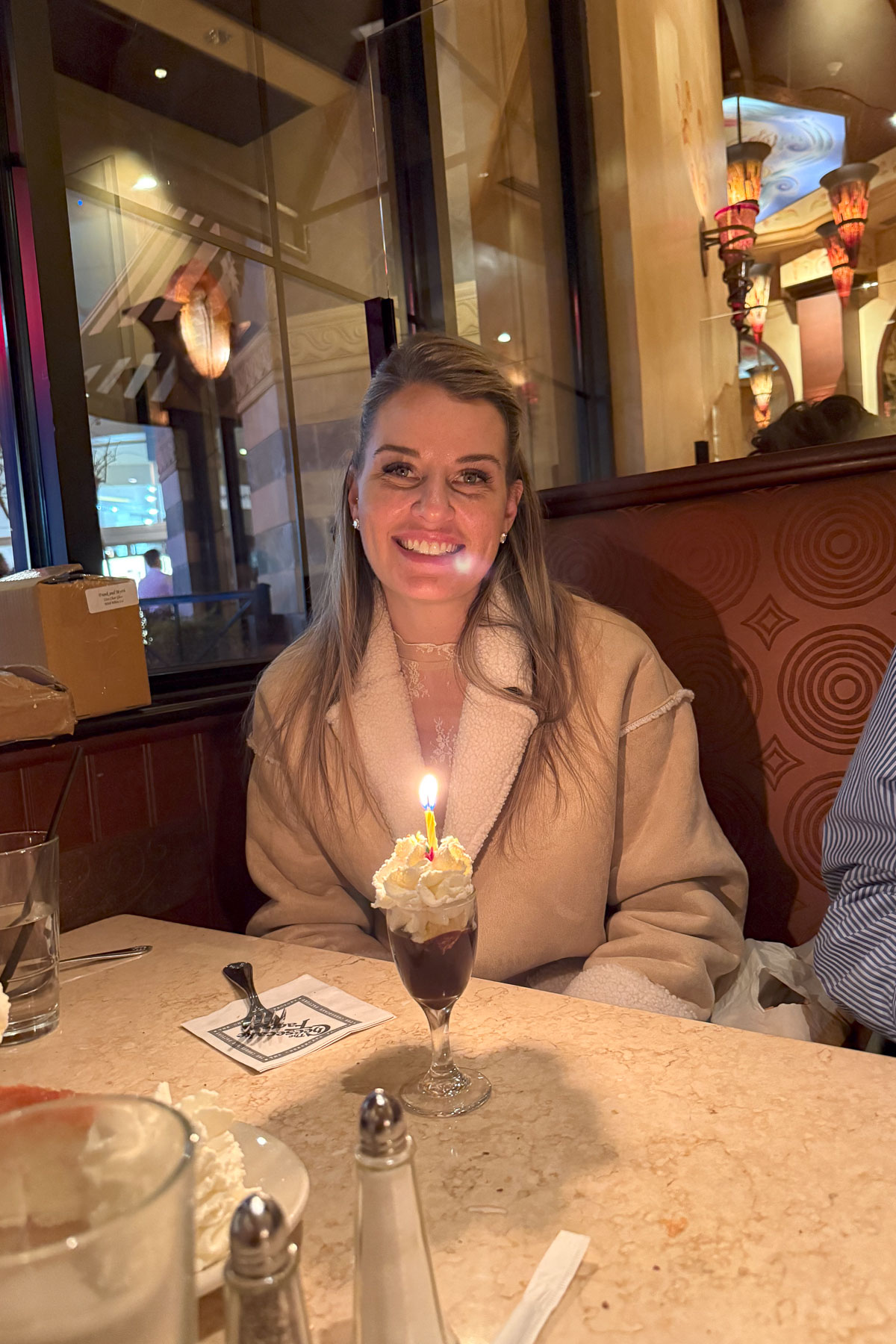 A woman smiling sits at a restaurant table with a small dessert topped with whipped cream and a lit candle in front of her, celebrating Sunday Sharing. The warmly lit interior, reminiscent of a cozy she shed, is adorned with ornate decorations.