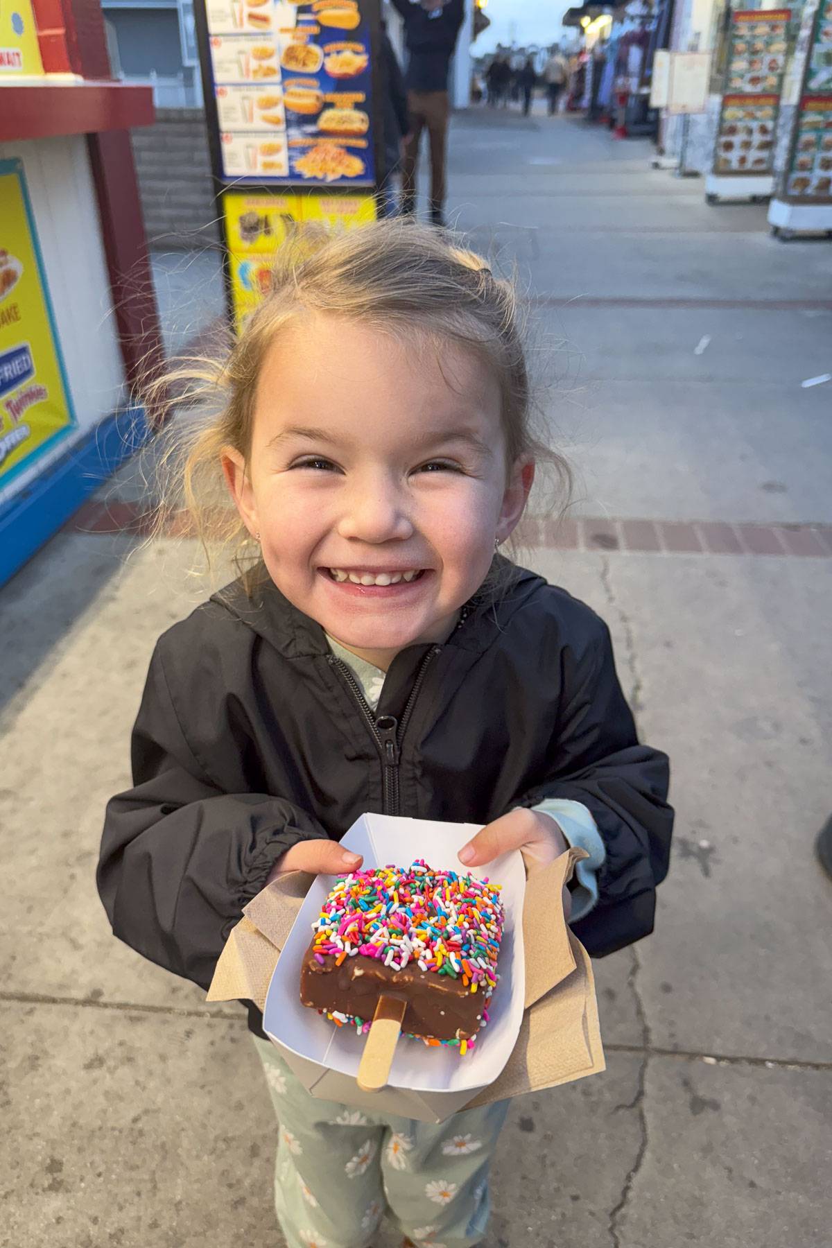 A smiling young girl in a black jacket holds a dessert on a stick, covered in chocolate and colorful sprinkles, on a busy street. The background features food stalls and a menu board.