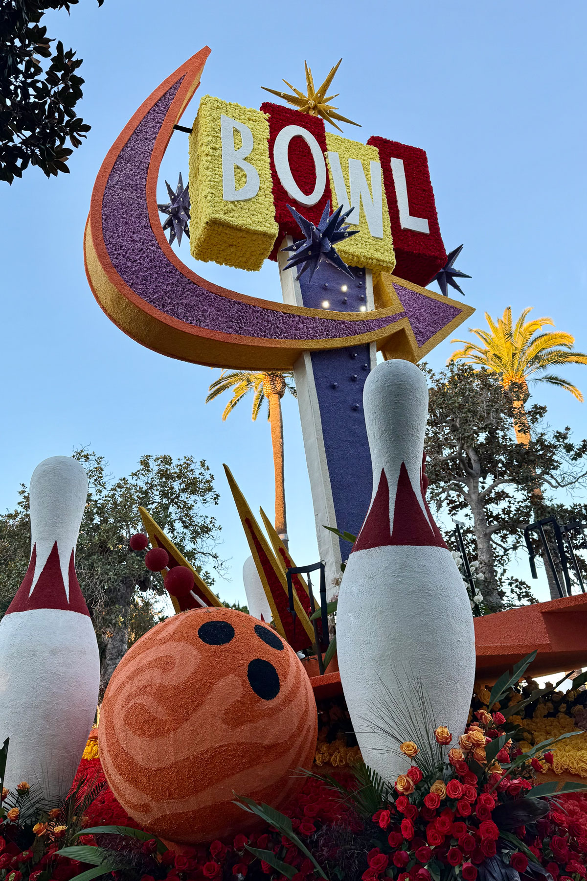 A colorful float featuring large 3D bowling pins and an orange bowling ball adorned with swirling patterns. Above, a vintage-style sign reads "BOWL" with stars and bright colors. Palm trees and a clear blue sky form the backdrop.