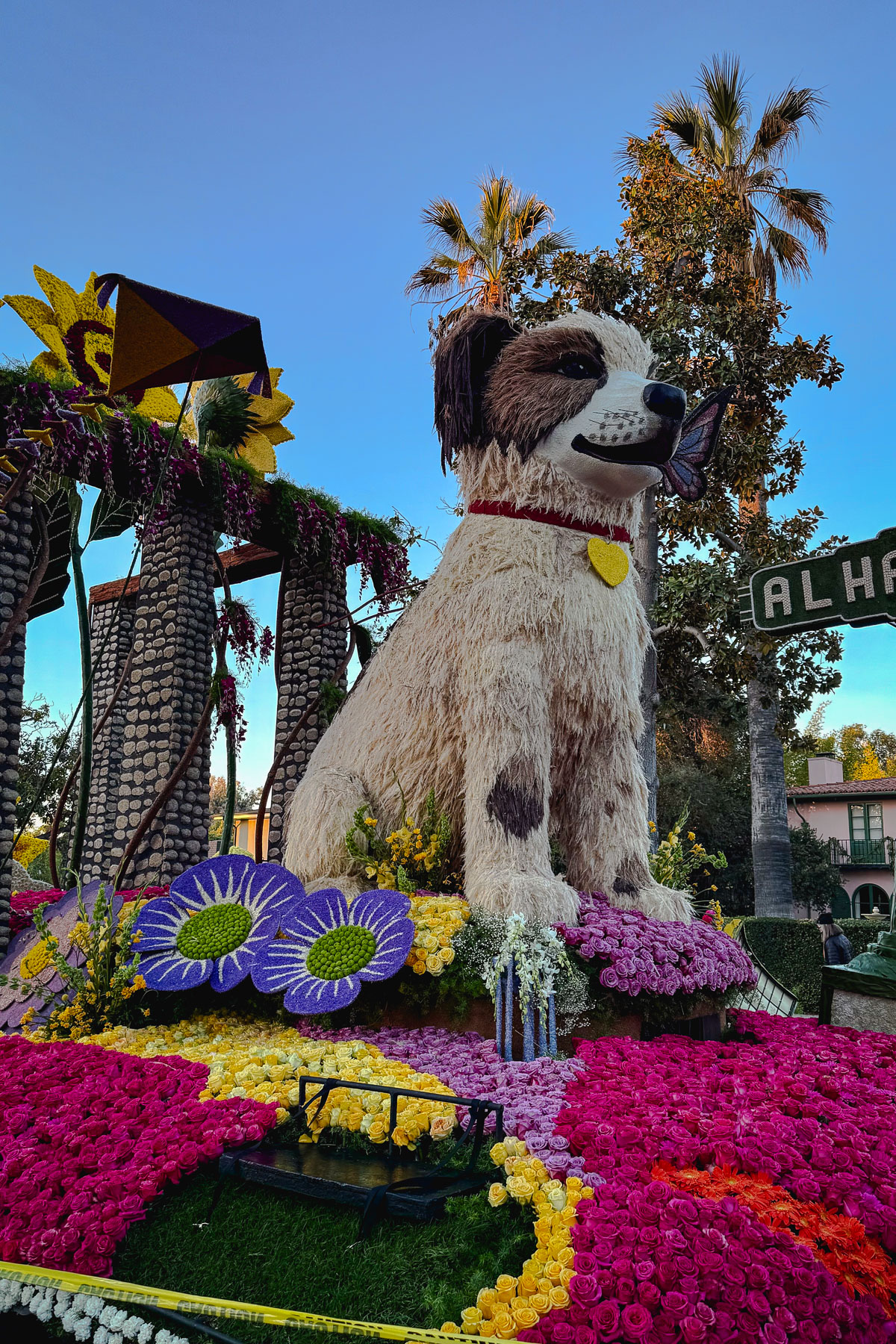 A parade float featuring a large dog sculpture made of flowers, surrounded by sunflowers and colorful floral arrangements. In the background, palm trees and a street sign are visible. The sky is clear and blue.