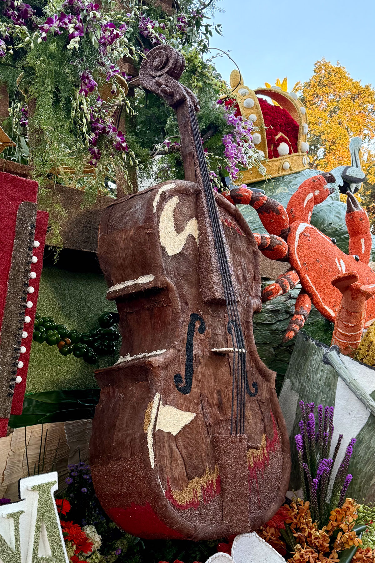 A large, decorative cello made from natural materials is part of a parade float adorned with flowers and plants. The background includes a bold, red crab sculpture and foliage, adding a festive and artistic atmosphere.